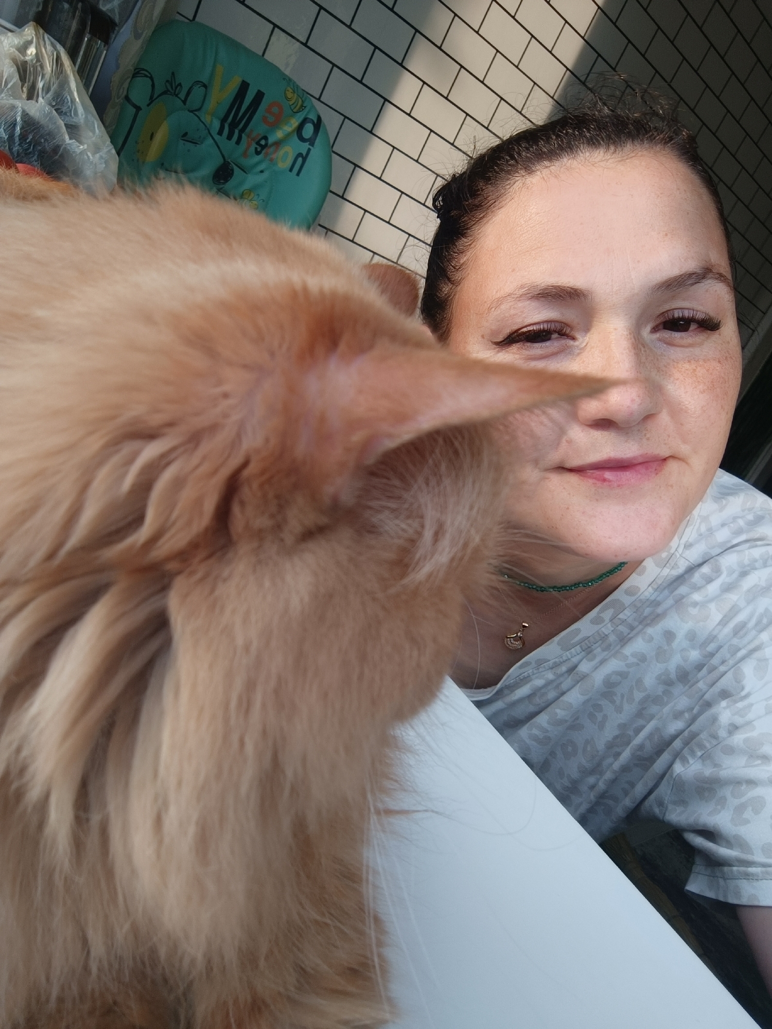 In the foreground of the image, a fluffy, light orange cat's fur obscures the left side of the frame. A woman with her hair pulled back in a bun is positioned to the right of the cat, looking towards the camera. She has fair skin with freckles, dark eyebrows, and noticeable eyelashes. She is wearing a light-colored t-shirt with a subtle leopard-print pattern. Around her neck, she wears a delicate green beaded necklace and a thin chain with a small, bird-shaped pendant. The background consists of white subway tiles and a partial view of what appears to be a blue, bear-shaped pillow with lettering. The scene is lit by natural light, casting shadows on the tiled wall. The context of Chaikovsky, Russia, is not evident from the visual elements of the image.