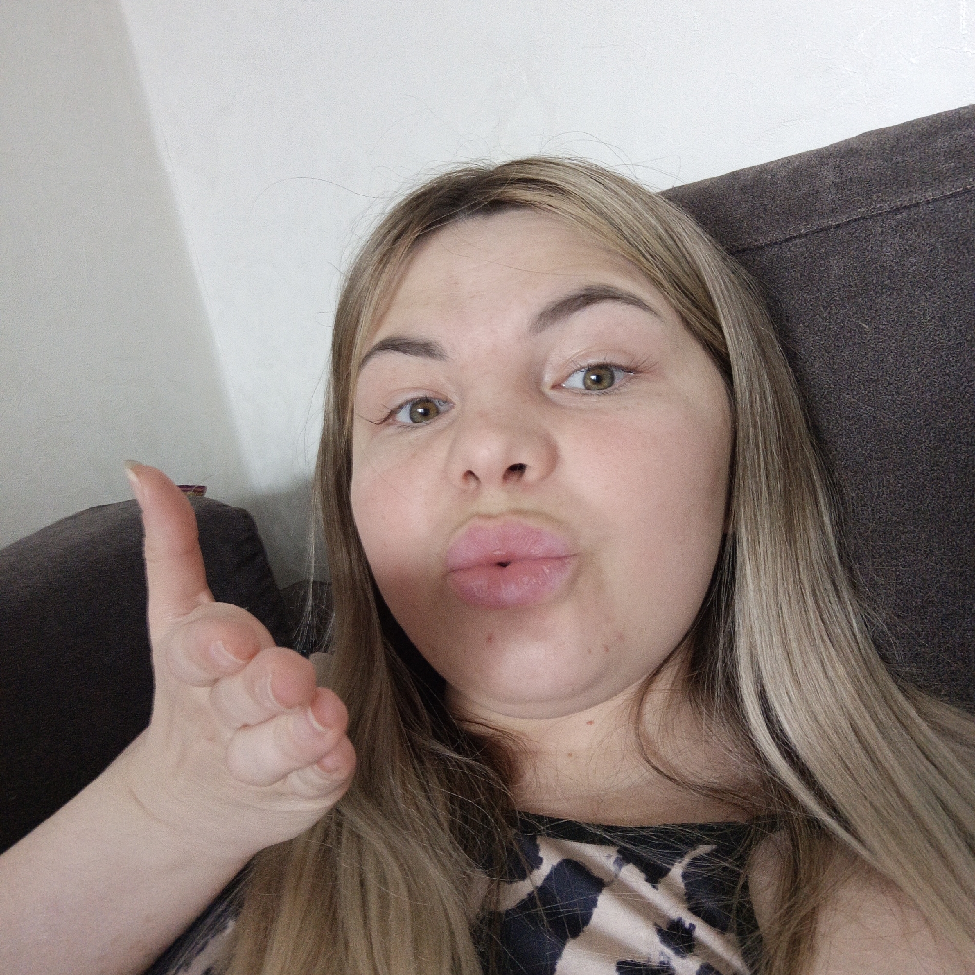 A woman is posing for a selfie. She is fair-skinned with light brown hair that is highlighted blonde. Her eyes are brown, and her lips are pursed into a kiss. She is holding her hand out, palm up, with her fingers curled. She is wearing a black and tan animal print top. She is sitting on a brown couch in front of a white wall. Location is Unknown, Unknown.