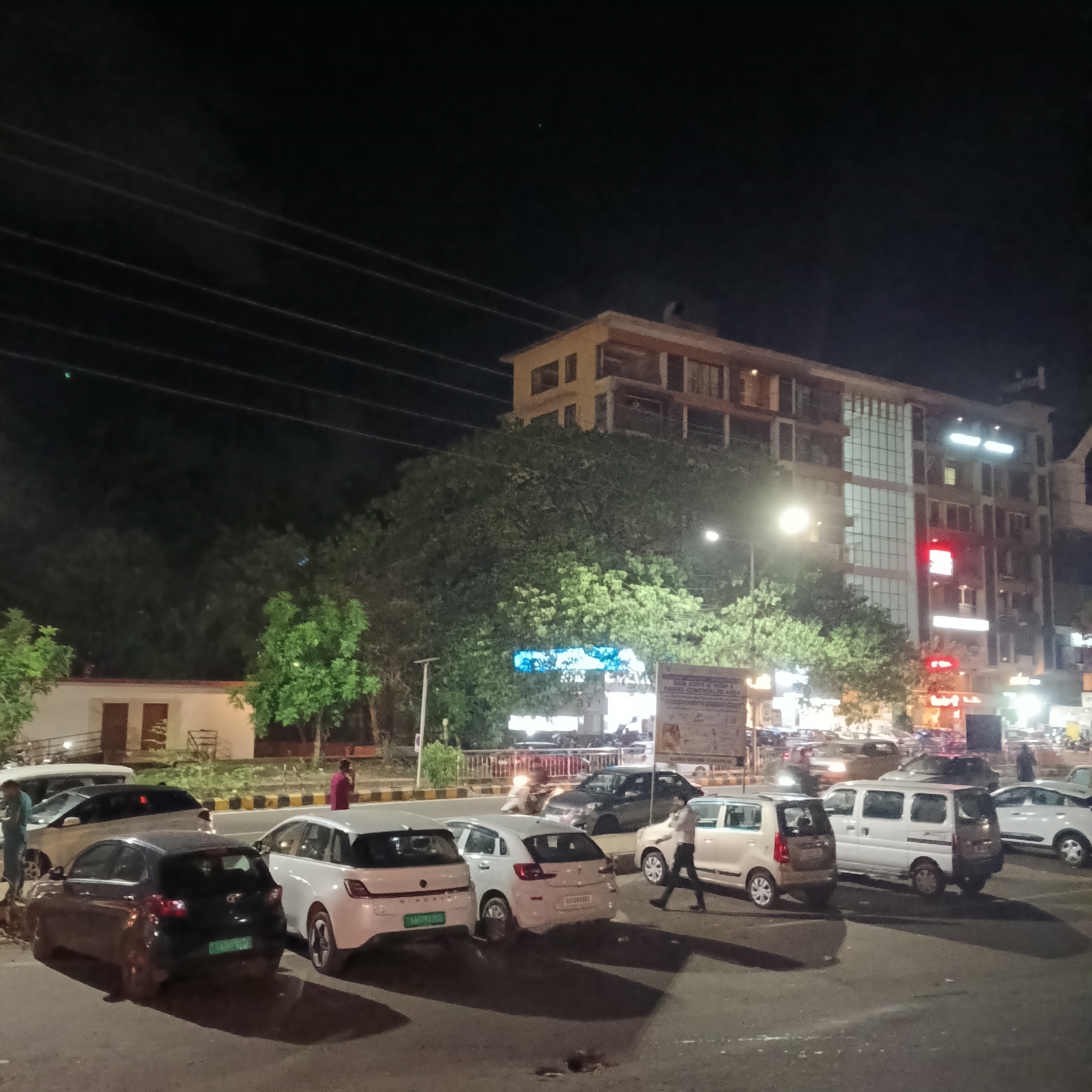 At night in Margao, India, the scene captures a busy street. Several cars are parked or moving. A person is walking between the parked vehicles. In the background, an illuminated building is visible with a large tree partially obscuring the view. Streetlights illuminate the scene. The sky is dark with visible electrical wires above.