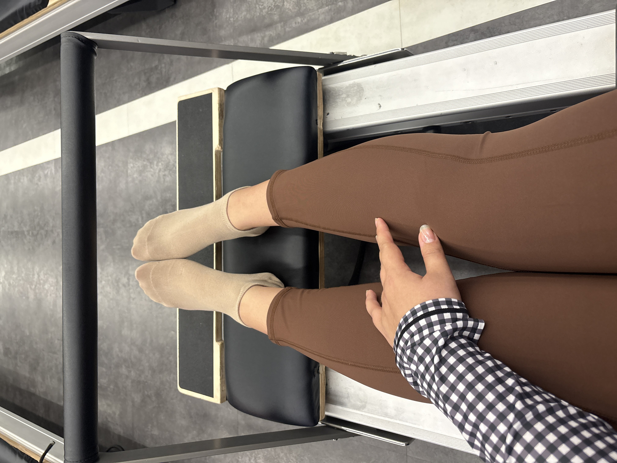 A close-up, overhead shot shows a person's legs and arm on a Pilates reformer. The subject is wearing brown leggings and light-colored socks. Their left leg is bent, with the foot resting on a black padded surface. The right leg is extended, and the hand with a checkered sleeve is resting on the thigh. The background appears to be a Pilates studio with a grey, textured floor and a portion of the Pilates reformer, which has metal tracks and a black carriage. The lighting suggests an indoor environment.