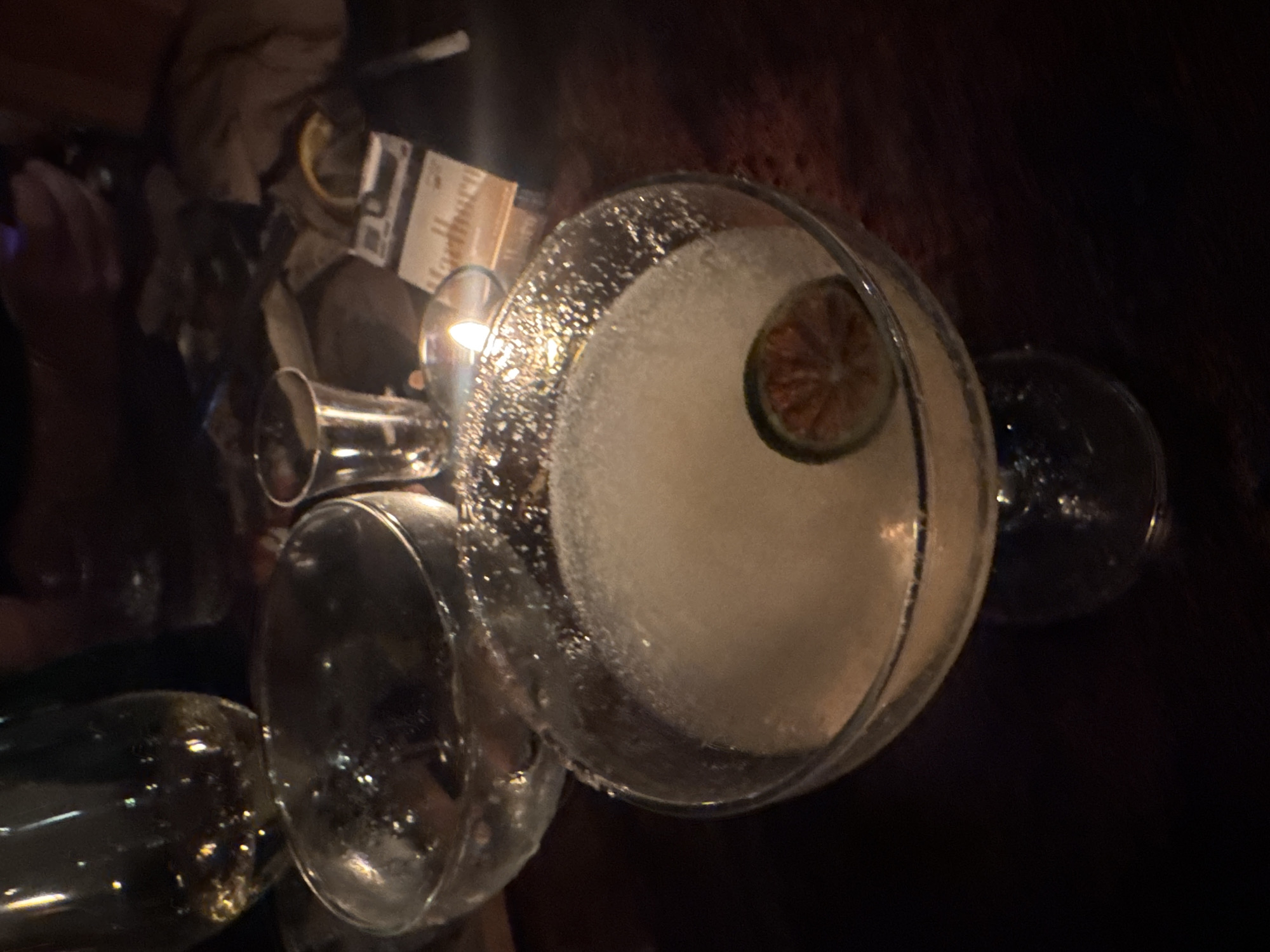 The image shows a cocktail on a dark wooden table with other glasses and items. The cocktail, in a large glass with a sugared rim, contains a white liquid and a lime wedge. A jigger and other empty glasses are present on the table. Based on the dim lighting and setting, it is likely taken at a bar or restaurant. There are no visible indications of the location being Kuta Selatan, Indonesia, in the image. Therefore, determining the location in the city without any evidence is impossible.