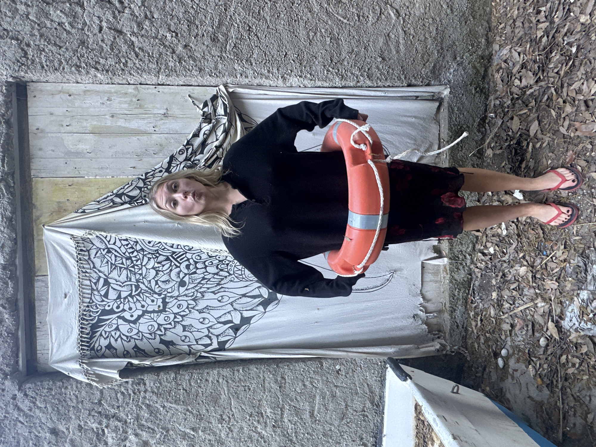 A fair-skinned woman stands in an alleyway, clutching a red and white life preserver. She wears a long-sleeved black shirt, a floral-patterned skirt, and red flip-flops. The alley is enclosed by rough, gray concrete walls. To her left is a weathered wooden door with a partially torn, white fabric covering it, decorated with a black and white floral pattern. To her right, the ground is covered with dry leaves and debris. The rear of a white and blue boat is visible at the bottom of the frame. The scene is likely located in the city of Lipari, Italy.