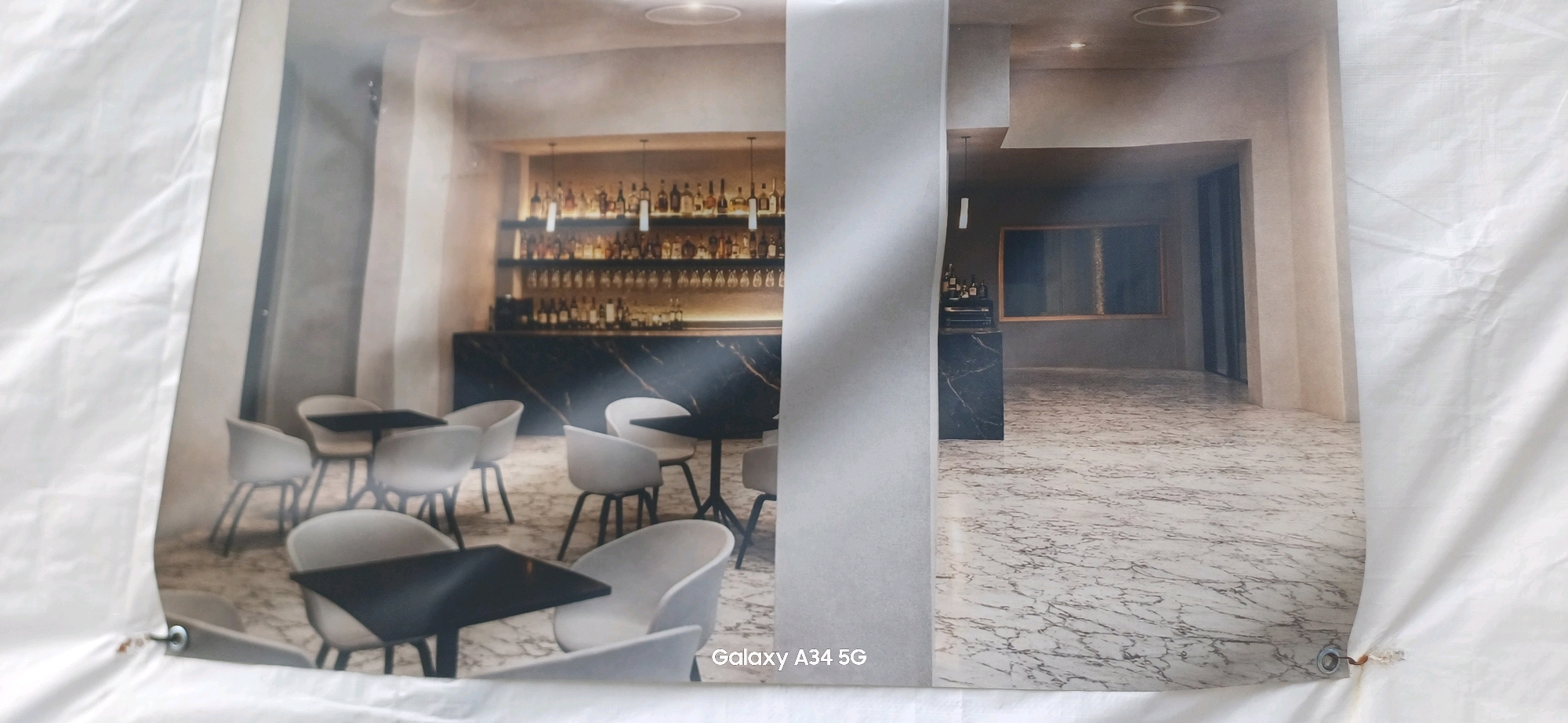 The image displays a photograph of an interior scene, likely a restaurant or bar, with a modern and sophisticated aesthetic. The left side of the photograph features a dining area with several white chairs and black tables. Behind the dining area is a bar with a marble countertop and numerous bottles displayed on shelves illuminated by warm lighting. The right side of the photograph shows a continuation of the interior with more marble flooring and a large framed artwork on a grey wall. The overall impression is one of a well-designed, upscale establishment. The "Galaxy A34 5G" watermark suggests the photo was taken with a Samsung phone. There are no people visible in the photograph. The location is not specified in the image itself, and there is no indication that it is in Abano Terme, Italy.