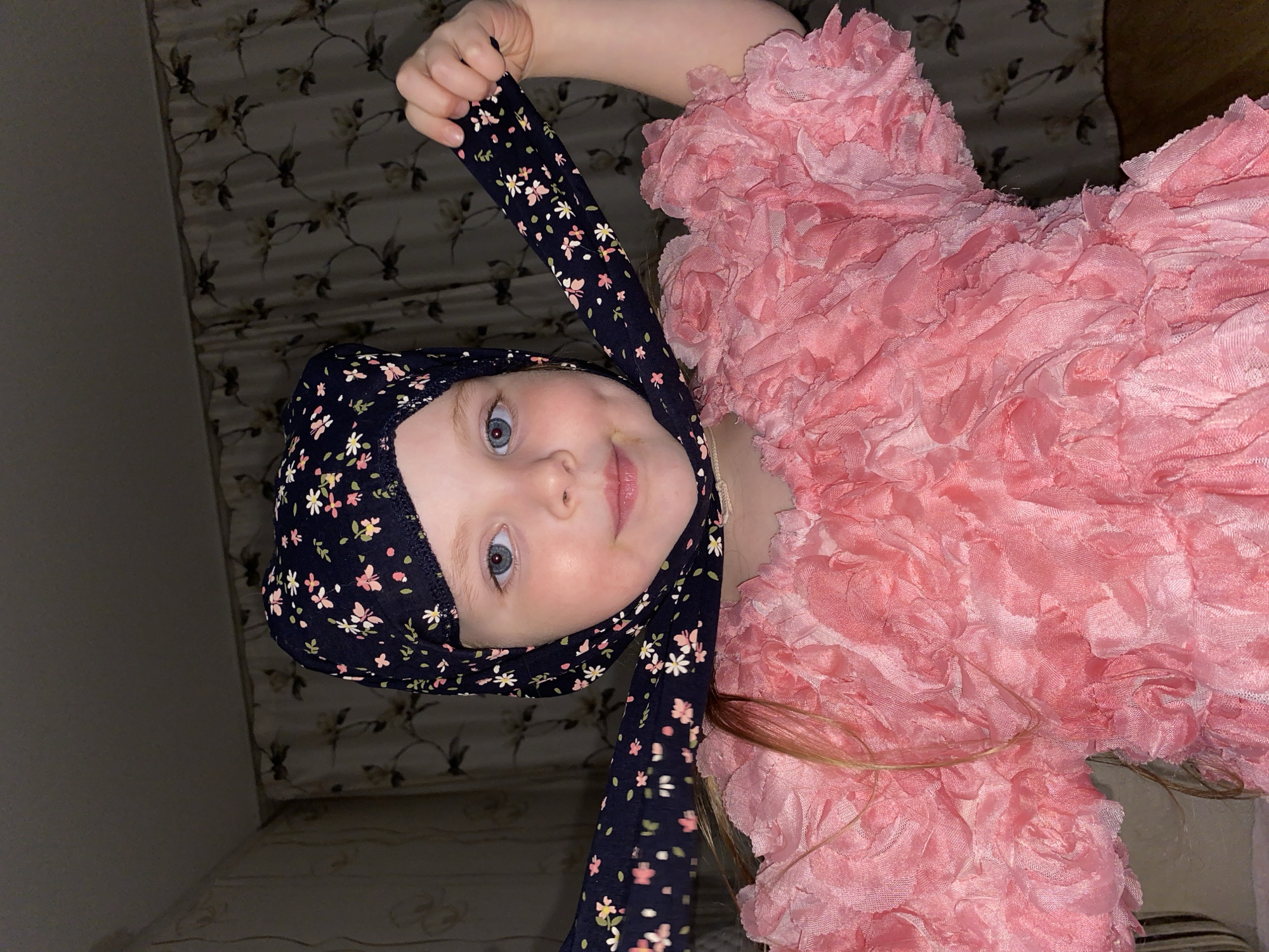 An indoor portrait features a young child, likely a girl, with fair skin, visible light hair strands, and striking blue eyes, looking directly at the camera with a gentle, slightly smiling expression. She appears to be lying down, with her head centered in the frame and her body extending towards the right.
The child is wearing a dark blue head covering, resembling a bonnet or bandana, adorned with a delicate, scattered pattern of small pink, white, and light green flowers. She playfully holds one of the long ties of this head covering with her raised right hand. Her attire also includes a vibrant pink top or dress, characterized by an intricate, ruffled texture that creates a dense, rosette-like appearance.
The background behind the child consists of a patterned curtain or tapestry featuring a subtle floral motif in muted tones of beige, brown, and dark grey-green. To the left of her head, a plain dark grey wall is visible. The setting suggests a comfortable home environment, possibly located in Votkinsk, Russia, given the provided context. The lighting is soft and indirect, contributing to an intimate and serene atmosphere. There is no visible text present in the image. The child's direct gaze and subtle smile convey a sense of innocence and engagement.