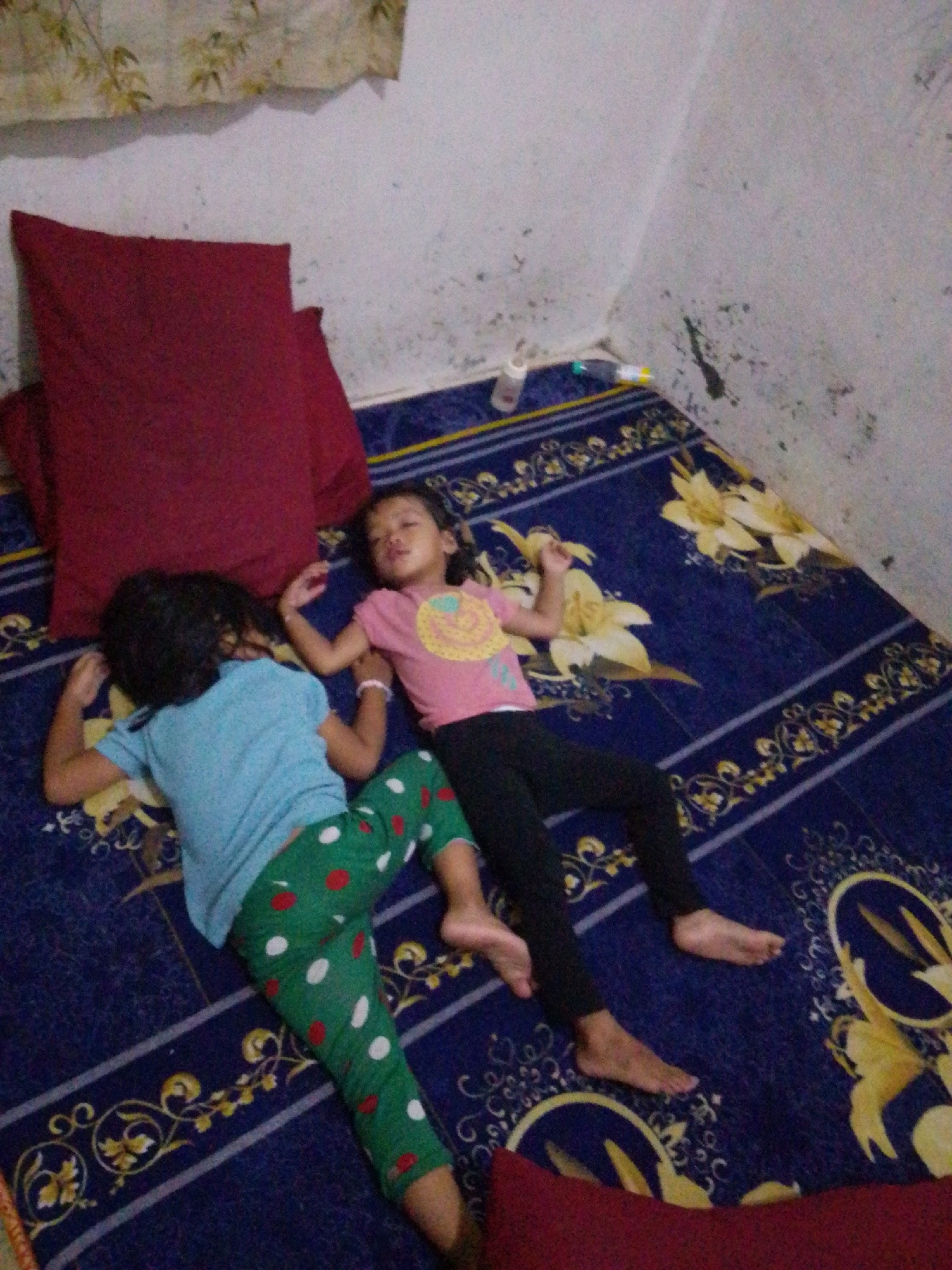 In an indoor setting, two young children are asleep on a blue mattress with a floral pattern. The child on the left is wearing a light blue t-shirt and green pants with red and white polka dots. Their head is turned away from the camera, and their arms are positioned as if they are sleeping. The child on the right is wearing a pink t-shirt with a yellow design and black leggings. This child's face is visible, and they appear to be sleeping soundly, with their arms resting by their sides. A large, dark red pillow is situated behind the child on the left. The background shows a light-colored wall, possibly painted, with some dark marks or stains. The lighting suggests it might be daytime, but the exact time is unclear. The overall scene depicts a quiet moment of rest.