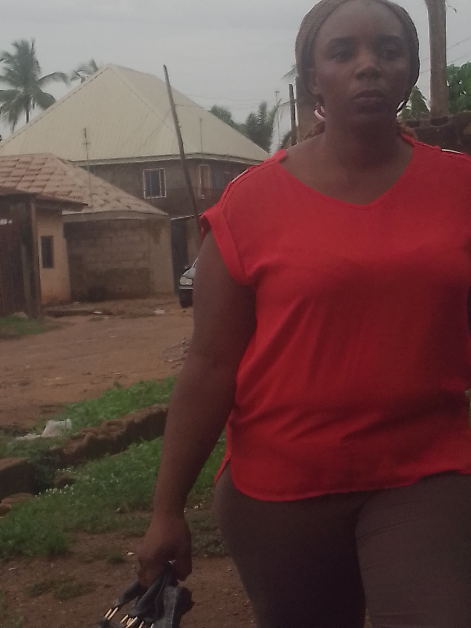 A woman in a red top and brown pants is walking on a dirt road in what appears to be a residential area. She is carrying a dark-colored bag in her left hand. Behind her are houses, including one with a distinctive pyramid-shaped roof. There are also trees, including a palm tree, visible in the background. A car is parked near one of the houses. The environment suggests a somewhat rural or developing urban area. The city is Unknown, Unknown.
