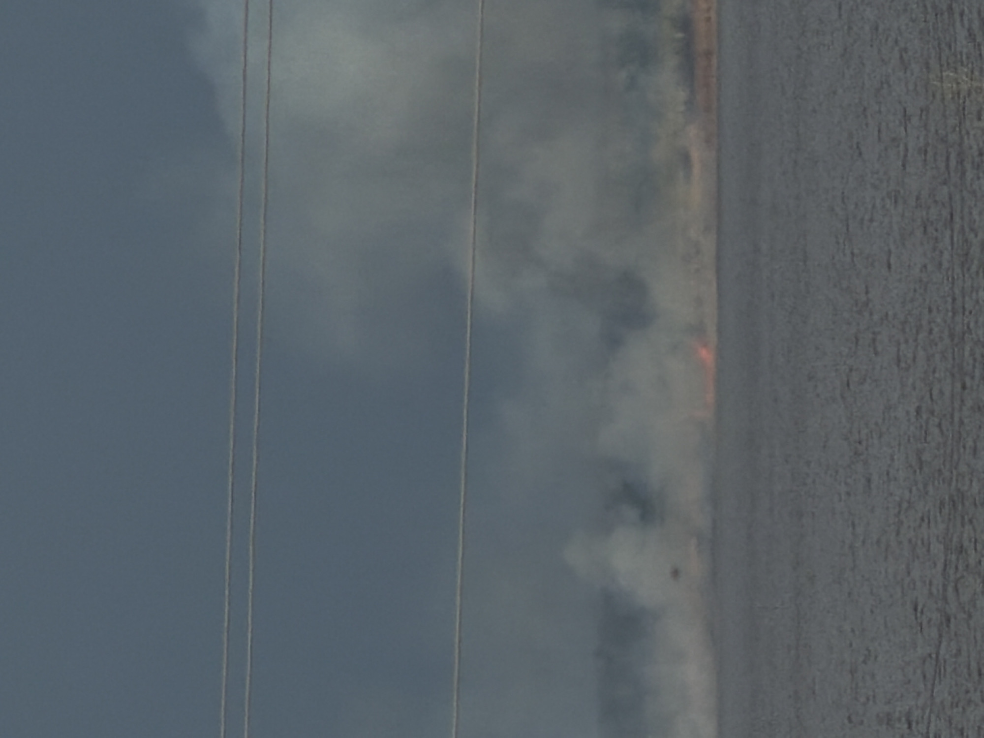 The image shows a landscape with a body of water in the foreground, possibly a river or lake. Above the water, there's smoke, suggesting a fire in the distance. The sky is overcast and gray. Power lines are visible in the foreground.
Without more specific details or context, it's impossible to confirm if this image is in Kamyshin, Russia.