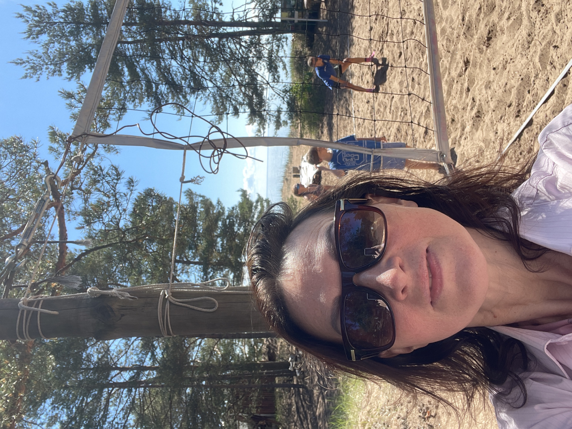 The image depicts an outdoor scene in Zelenaya Roshcha, Russia, under a clear blue sky. In the foreground, an adult female is taking a selfie, visible from the chest up. She has dark brown hair and wears large, dark-framed sunglasses, which reflect a handheld device and surrounding trees. Her attire consists of a light-colored, collared button-up shirt.
Behind her, a sandy beach volleyball court is visible, featuring a rustic net supported by a wooden pole on the left, secured with ropes. Three individuals are present on or near the court. On the far left, a female child in a dark top is partially visible near the net. Centrally, a male child wearing a blue t-shirt with a white graphic stands near the net. To the right, a male individual in a blue t-shirt, dark shorts, and bright pink athletic shoes is positioned in a defensive stance on the sand, ready to play volleyball.
The background features a dense pine forest lining the sandy area. Beyond the forest and a slight elevation of sand, a large body of water, likely a lake or sea, extends to the horizon. Small, weathered structures are faintly visible among the trees on the far left. The scene is brightly lit by direct sunlight, casting shadows on the sand.