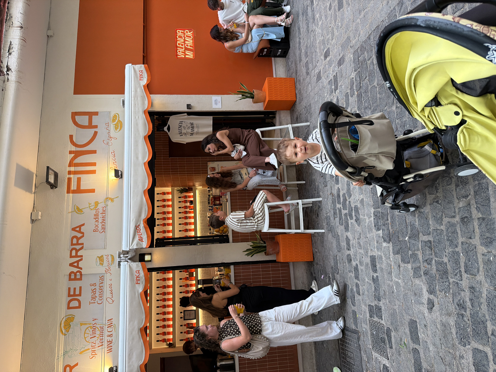 In an outdoor urban setting, a street scene unfolds with a food and drink establishment adorned with orange and white awnings and signage. The main facade displays text including "DE BARRA," "FINCA," "Spritz. Vinos y Vermut," "Tapas & Conservas," "Bocadillos Sandwiches," and "Quesos y Ibericos." Behind the counter, rows of red bottles, likely aperitif, are illuminated. Several people are visible; a woman in a polka dot top and white pants holds a drink and a bag, standing near a woman in a black outfit pouring a beverage. A young boy in a striped shirt sits on a stool, looking towards the camera. Another group of individuals are seated on stools and benches. To the right, a neon sign reads "VALENCIA MI AMOR." The ground is paved with cobblestones. In the foreground, a child is seated in a stroller, and another stroller is partially visible. The overall atmosphere suggests a casual, social gathering at an establishment.