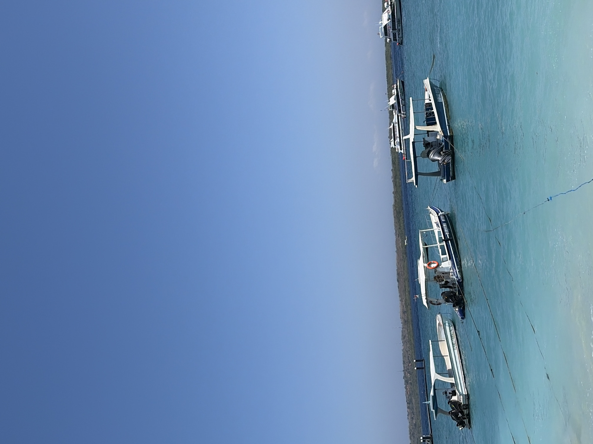 This image shows a body of turquoise water with several boats. In the foreground, a white boat with green trim is visible. To its right is a blue and white boat with a canopy and an orange life preserver. In the background, there are larger white passenger boats. The scene is set against a clear blue sky and a distant coastline with vegetation and structures. The location is likely near Nusa Penida, Indonesia, a popular tourist destination known for its clear waters and boat excursions. The boats appear to be anchored, suggesting a calm day for water activities. There are no people clearly visible in the image.