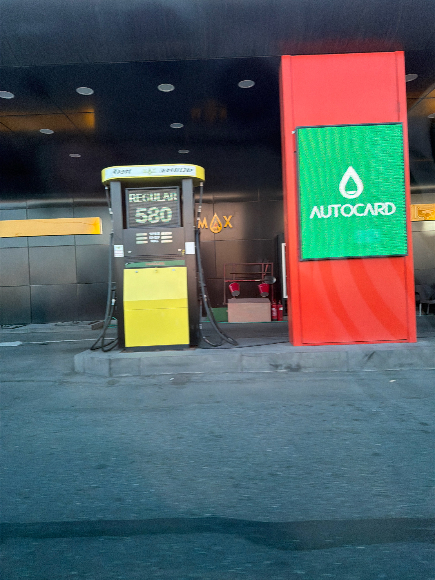 The image shows a gas station in Yerevan, Armenia. The gas station has a prominent red pillar with a large green LED screen displaying the "AUTOCARD" logo and a droplet symbol. To the left of the red pillar is a yellow and black gas pump displaying "REGULAR 580". The background features a dark ceiling with circular lights and a dark tiled wall. A yellow rectangular sign is visible on the left wall. The ground in front of the gas station is paved. The scene appears to be in an outdoor setting, possibly at dusk or night, given the lighting. There are no people clearly visible in the image.