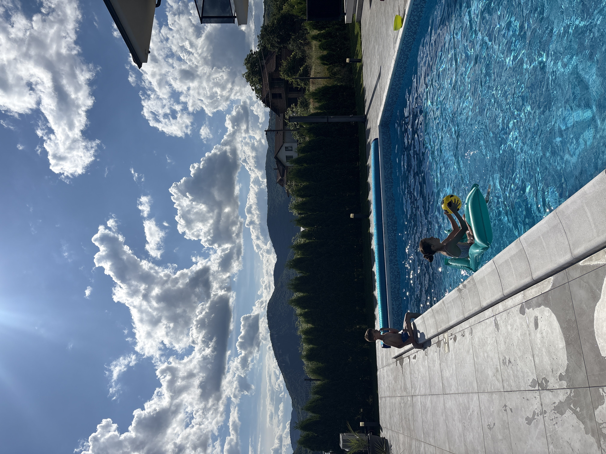 A boy sits on the edge of a swimming pool, his legs dangling over the water. A girl floats in the pool on an inflatable raft, holding a yellow ball. The swimming pool is surrounded by a tiled patio and a hedge. In the background, there are hills and houses under a cloudy sky.