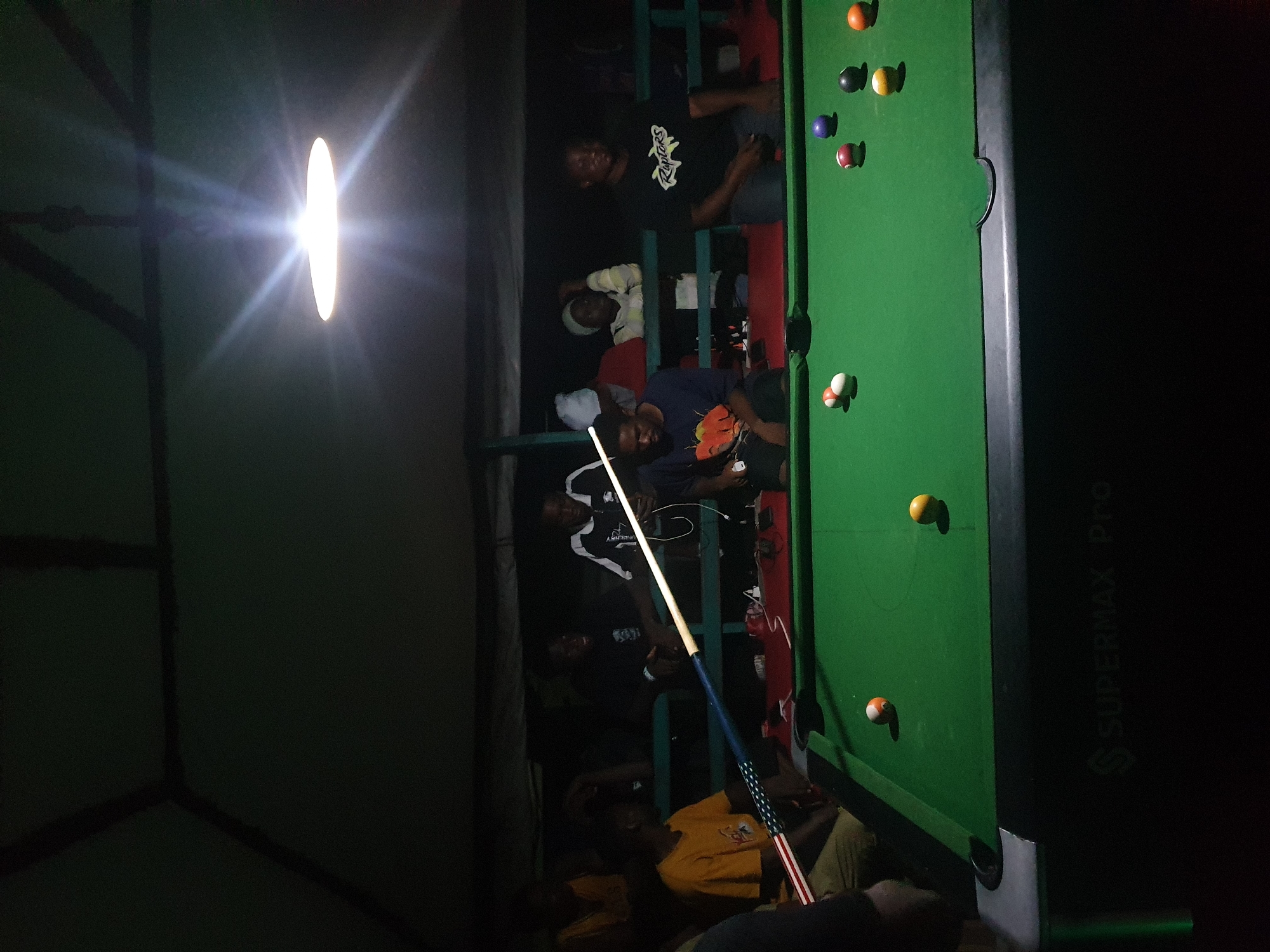 The image depicts an indoor pool hall, likely in Lokoja, Nigeria, where a group of young men are engaged in a game of pool. The setting appears to be a casual, possibly outdoor or semi-outdoor venue, suggested by the visible structure of the roof and the dim, atmospheric lighting provided by a single overhead lamp. The pool table, covered in green felt, is the central focus, with several billiard balls scattered across its surface, including a cue ball. A pool cue, adorned with a distinctive American flag pattern, is held by one of the players. The individuals are casually dressed, some observing the game, while others are actively participating or preparing to play. The overall atmosphere suggests a relaxed social gathering centered around the game of pool.