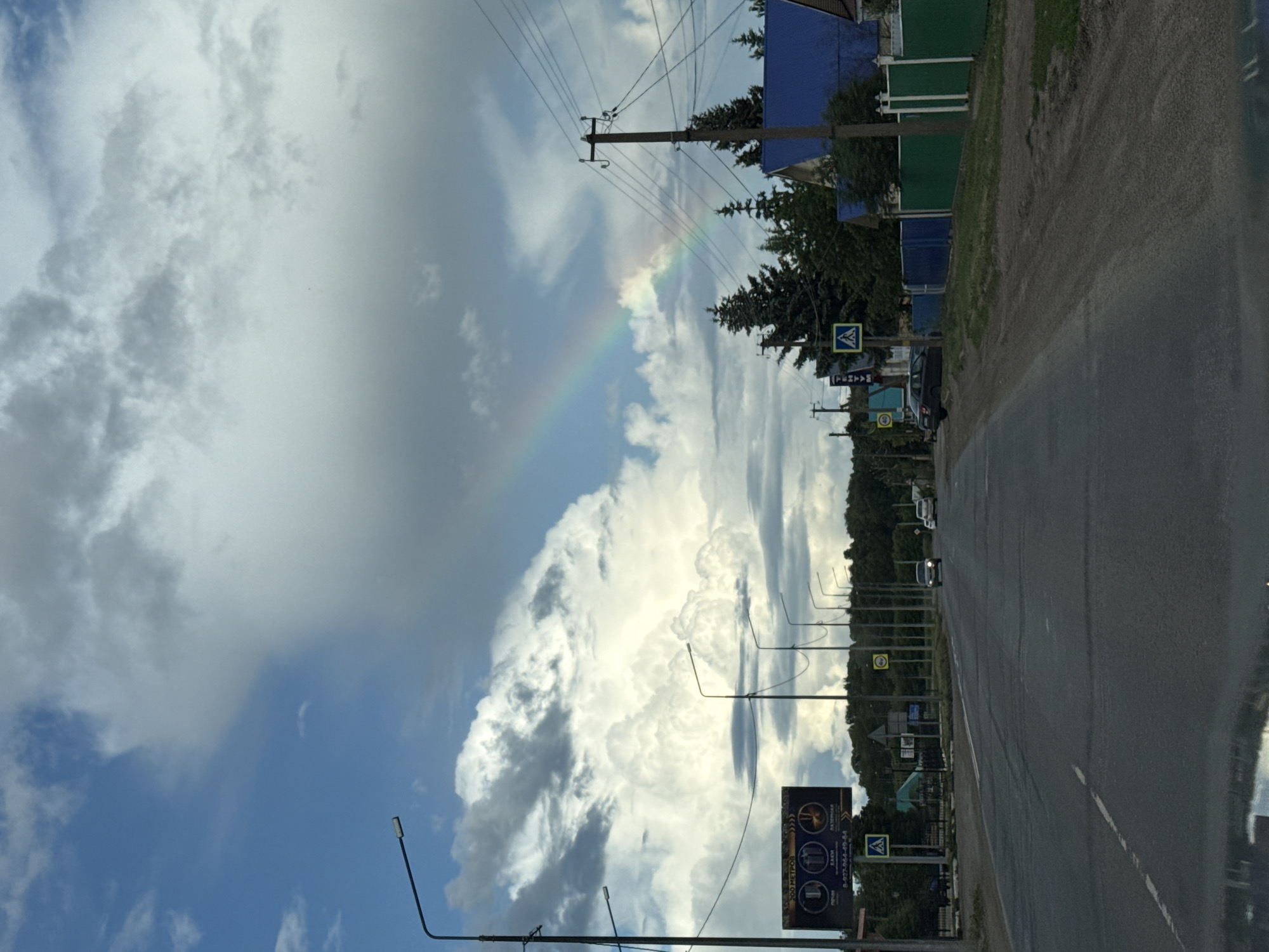 The image captures a daytime scene on a rural road in Нуркеево, Russia, under a partially cloudy sky with a visible rainbow. The perspective is from inside a vehicle, indicated by a reflection at the bottom edge.
The sky occupies the upper two-thirds of the frame, displaying a mix of deep blue sky on the left and varying densities of white and grey cumulus clouds across the expanse. A faint but distinct rainbow arc is present in the upper right quadrant of the sky.
The asphalt road, which appears worn with cracks and patches, extends from the foreground into the distance, curving slightly to the right. A faded white solid line marks the left edge of the carriageway. Two light-colored passenger vehicles are visible in the distance, traveling away from the viewer on the road.
On the left side of the road, a series of tall black streetlamp poles with horizontal light fixtures line the route. Further along this side, a large rectangular billboard is prominent, displaying an advertisement with Cyrillic text, including "200 МЕТРОВ" (200 METERS) and a phone number "8-927-964-49-64," alongside three circular icons depicting what appear to be services like a wrench, a battery, and a spark plug or tire. Below the billboard, a yellow triangular road sign with a black pedestrian crossing symbol is visible.
On the right side of the road, a dirt and gravel shoulder transitions to grassy verges. Several utility poles with multiple overhead power lines are present. A blue-roofed house is visible behind the utility lines, surrounded by coniferous trees. Further down the road on the right, another yellow triangular pedestrian crossing sign is seen near a blue and green segmented fence. A circular yellow speed limit sign displaying "40" km/h is also visible on this side. Dense green foliage and trees line both sides of the road, forming a horizon line in the middle distance. No people are visible.