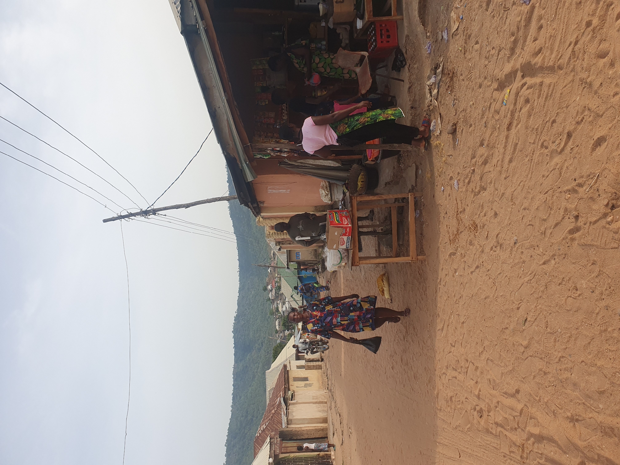 The image depicts a vibrant outdoor scene in Lokoja, Nigeria, likely along a rural or semi-urban roadside. The ground is unpaved, consisting of reddish-brown dirt or sand with numerous footprints and some litter scattered about.
In the foreground, a woman wearing a colorful, patterned dress stands on the dirt road, holding a black object, possibly a bag or container, in her right hand. She is looking towards the viewer with a slight smile. Behind her, along the left side of the frame, several single-story buildings with corrugated iron roofs line the road.
To the right, under a makeshift awning, is a small shop or stall. Several people are gathered here. Two women are seated; one in a pink top and green patterned skirt, and another in a green patterned dress. They appear to be resting or socializing. Various goods and household items are displayed on shelves and tables under the awning, including red plastic crates. A man is standing next to a wooden table holding a cardboard box that clearly displays the brand "INDOMIE" (likely instant noodles). Another person is partially visible behind the seated women. Further down the street, more buildings and what appears to be a motorcycle are visible.
In the background, a range of green, tree-covered hills or mountains stretches across the horizon under an overcast sky. A utility pole with multiple power lines runs vertically on the left side of the image, extending into the sky, suggesting electrical infrastructure in the area. The scene captures daily life with people engaged in commerce and routine activities under natural lighting conditions.
