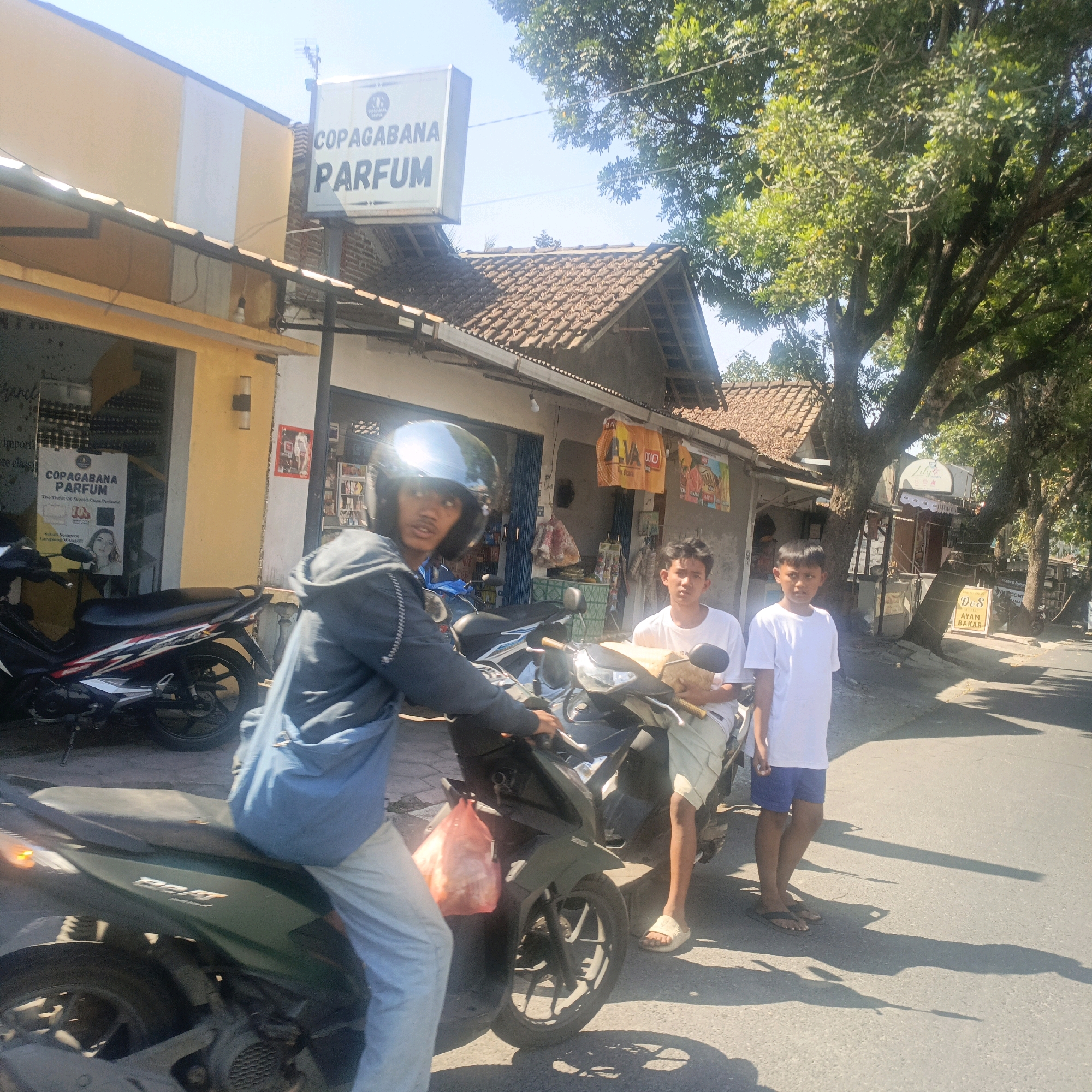 In Salatiga, Indonesia, on a bright, sunny day, a street scene unfolds. In the foreground, a young man wearing a dark helmet and a grey hooded jacket is seated on a dark green scooter. He is looking towards the camera. To his right, another young man in a white t-shirt and light-colored shorts stands next to a beige scooter, holding a box. Behind them, a third young man in a white t-shirt and blue shorts stands on the road. In the background, a building with a yellow facade and a tiled roof is visible, featuring a sign that reads "COPAGABANA PARFUM". Several trees with dense green foliage provide shade along the street, and power lines are strung overhead. The road surface is asphalt, with shadows cast by the sun. A red plastic bag is hanging from the side of the scooter in the foreground.