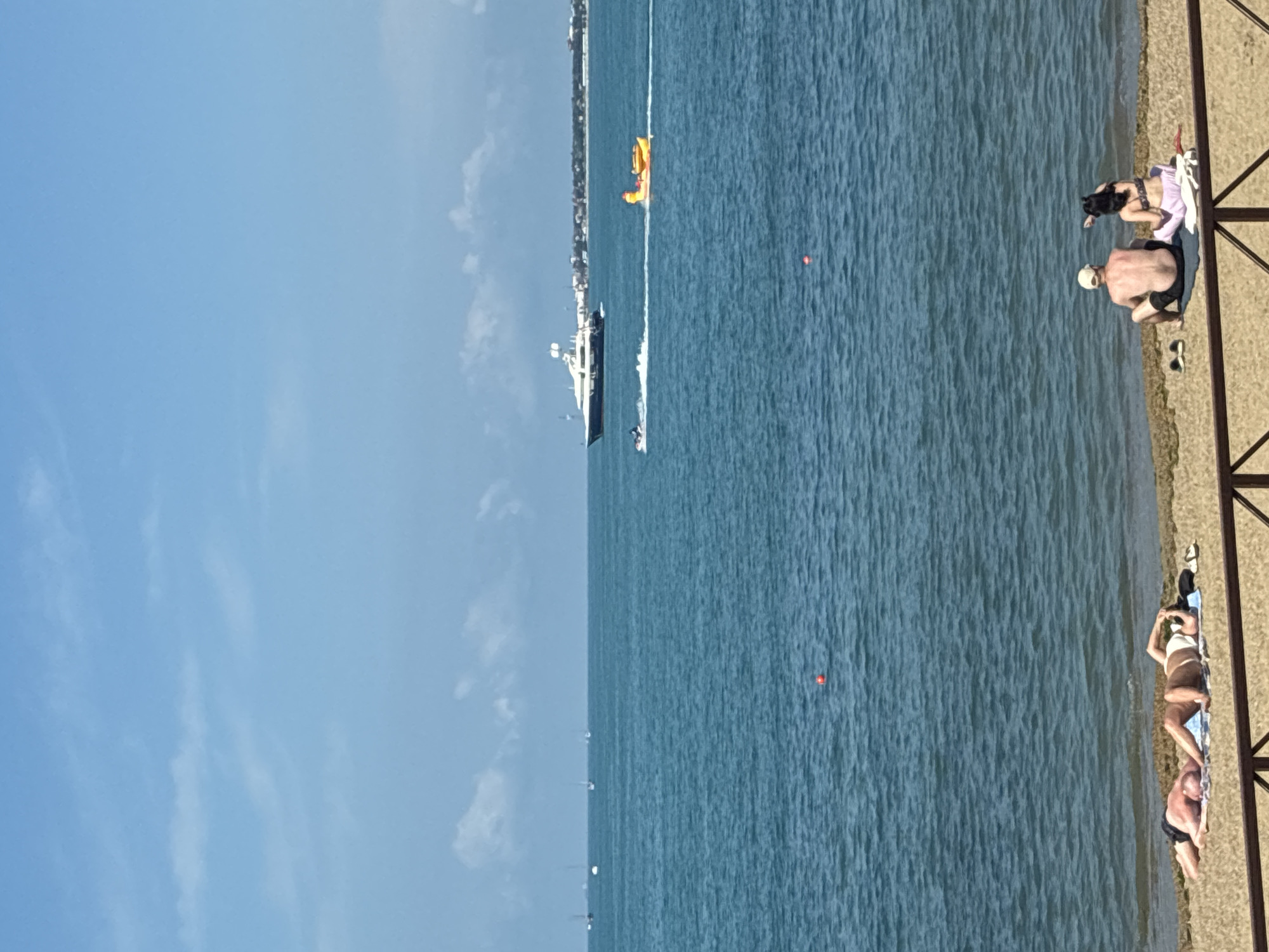 In Gelendzhik, Russia, on a sunny day with a clear blue sky and scattered clouds, a tranquil scene unfolds at the Black Sea. The foreground features a sandy beach where several individuals are relaxing. On the left, two people are lying on towels, sunbathing. Further to the right, a couple sits on the sand, facing the sea, with one person pointing towards the water.
The sea itself is a vibrant blue-green, with gentle waves lapping the shore. Out on the water, a large white yacht sails in the distance, accompanied by a jet ski leaving a white wake. A yellow inflatable towable tube, shaped like a duck, is being pulled behind the jet ski. Several other sailboats are also visible on the horizon, along with the distant coastline of Gelendzhik, characterized by buildings and greenery. A small orange buoy floats in the middle of the sea. A wooden fence runs along the bottom of the frame, separating the beach from an unseen area.