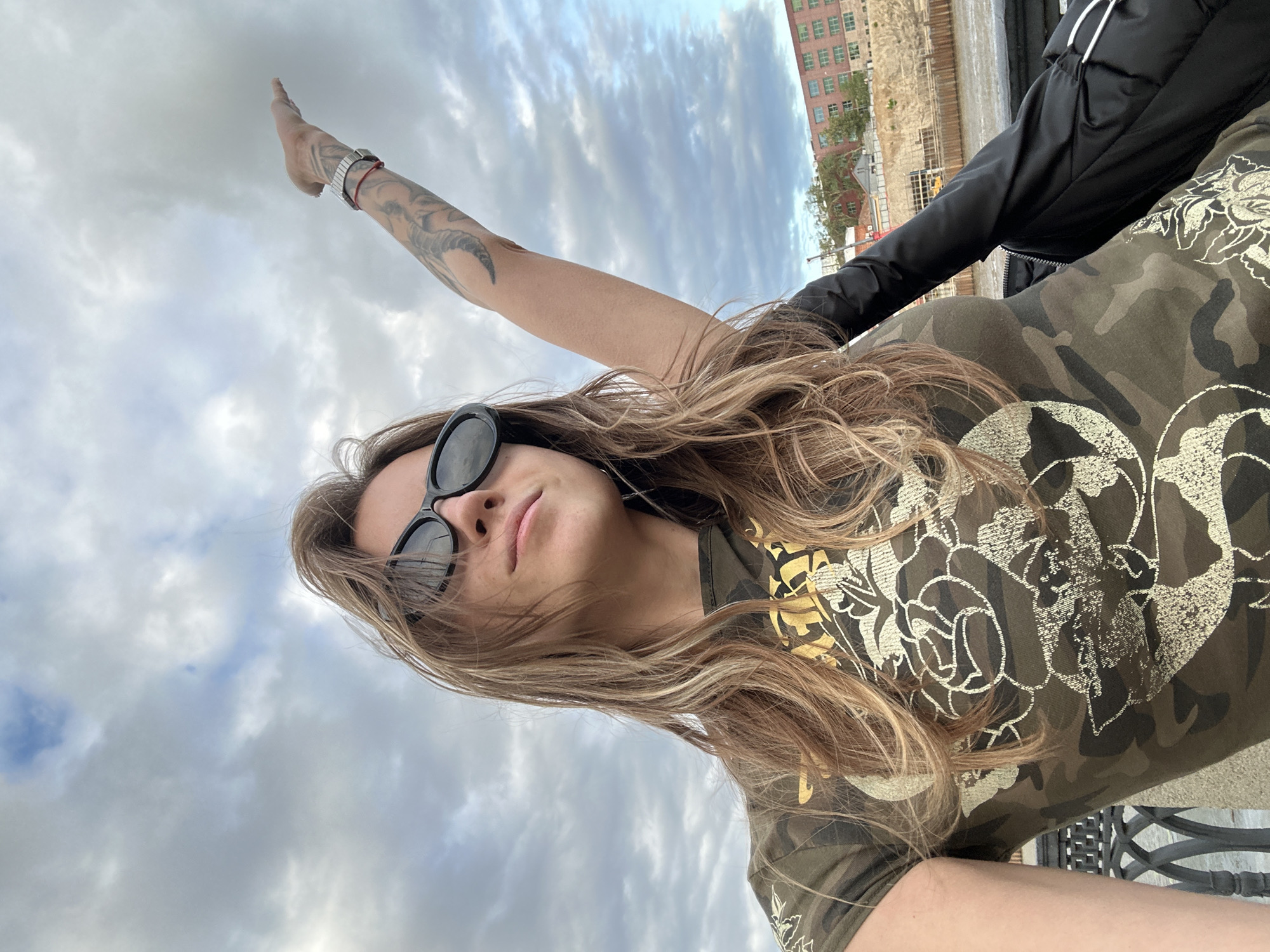 A person is captured in a low-angle, close-up shot, with their face and upper body dominating the frame. They are wearing sunglasses and a camouflage t-shirt with a white floral design. Their right arm is raised, with visible tattoos on their forearm, and their fingers are pointed upwards. The background consists of a cloudy sky and hints of an urban environment, including a building and some construction. The overall impression is that of a selfie taken outdoors, possibly in Moscow, Russia, given the location context. The lighting suggests it could be daytime, with overcast conditions. The person's pose and the upward-pointing gesture could imply a sense of aspiration or triumph.