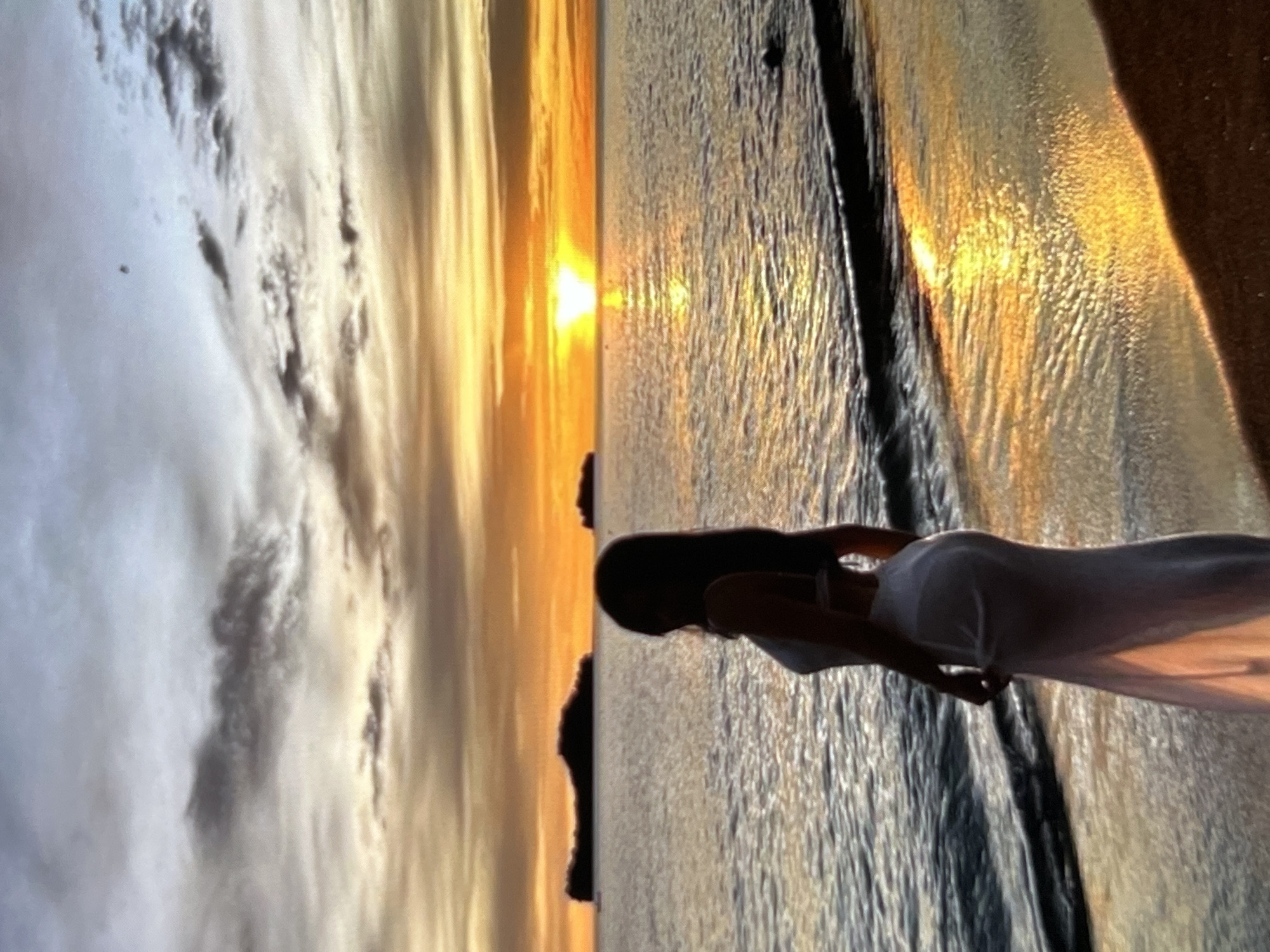 The image depicts a serene sunset scene at a beach. A person, silhouetted and wearing a white dress, stands near the water's edge, facing away from the camera and towards the ocean. The sun is setting on the horizon, casting a warm, golden glow across the water and sky. Islands are visible in the distance. The sand is wet from the receding tide, and small waves are gently lapping at the shore. The sky is a mix of orange, yellow, and grey clouds. The scene is tranquil and evocative of a peaceful evening by the sea. This image is not located in Montfermeil, France.