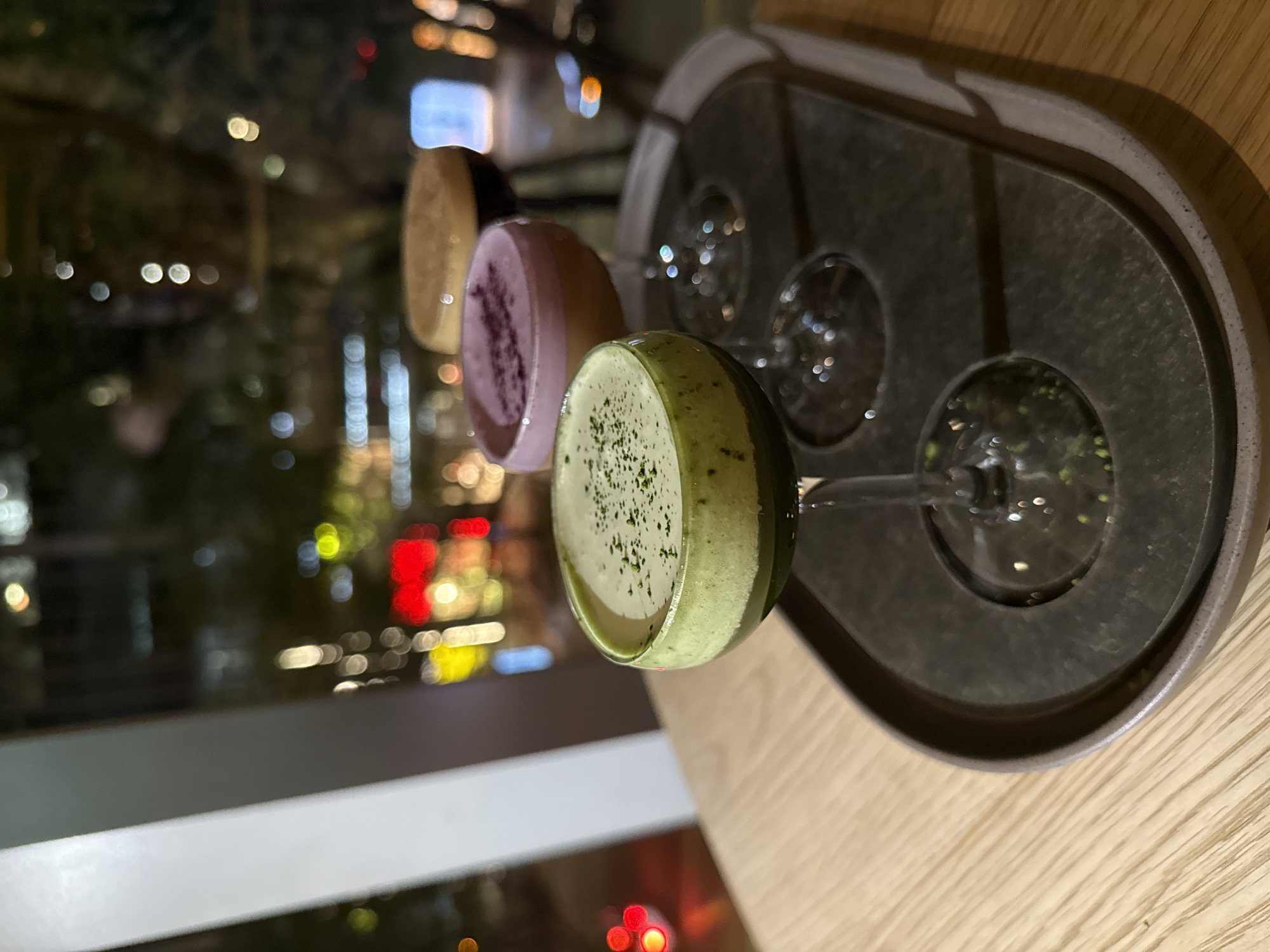 Here is a forensic description of the image:
The image depicts a tray with three stemmed glasses, each containing a different colored liquid, likely a cocktail or dessert, arranged on a wooden table near a window. The glass closest to the viewer holds a light green beverage, possibly matcha-flavored, topped with dark green sprinkles. The next glass has a purple-colored liquid, and the furthest glass is filled with a beige or light brown concoction. The tray appears to be made of a dark gray material, possibly metal or stone. The tray has four circular impressions indicating where glasses can be placed.
Outside the window, the blurred lights of a city skyline can be seen. The lights suggest it is nighttime. The city lights, especially the red hues, coupled with the blurred quality, suggest the location is an urban environment. Due to image analysis restrictions I cannot verify specific locations.