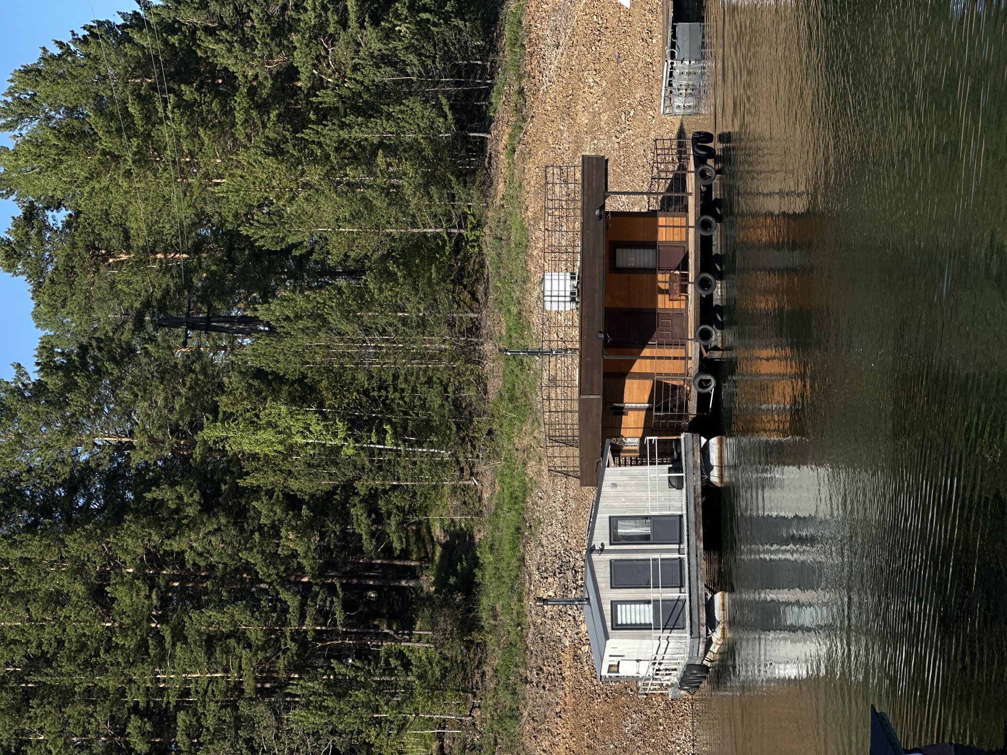 A serene waterfront scene features two moored structures alongside a body of water, bordered by a dense coniferous forest. The water, likely a lake or wide river, occupies the right side of the frame, its surface gently rippled and reflecting the sky and the structures.
On the left, a strip of rocky, gravelly ground extends from the water's edge, transitioning into a thick forest of tall, green coniferous trees. A clear blue sky is visible above the treetops in the upper left. Faint power lines run through the forest, supported by utility poles.
Two structures are prominent at the water's edge. The one on the right is a larger, multi-story wooden building or spacious houseboat with a warm brown exterior, featuring windows, doors, and a metal railing. Several black tires hang along its water-facing side, likely acting as fenders. To its left, partially obscured, is a smaller, more modern-looking gray houseboat or floating cabin, characterized by black-framed windows, a dark roof with a chimney/vent pipe, and white railings. A white container or tank is visible near the wooden structure.
The scene appears calm and undisturbed, with no people or specific activities taking place. Bright daylight suggests it is daytime, possibly morning or afternoon, under clear weather conditions. No visible text is present.