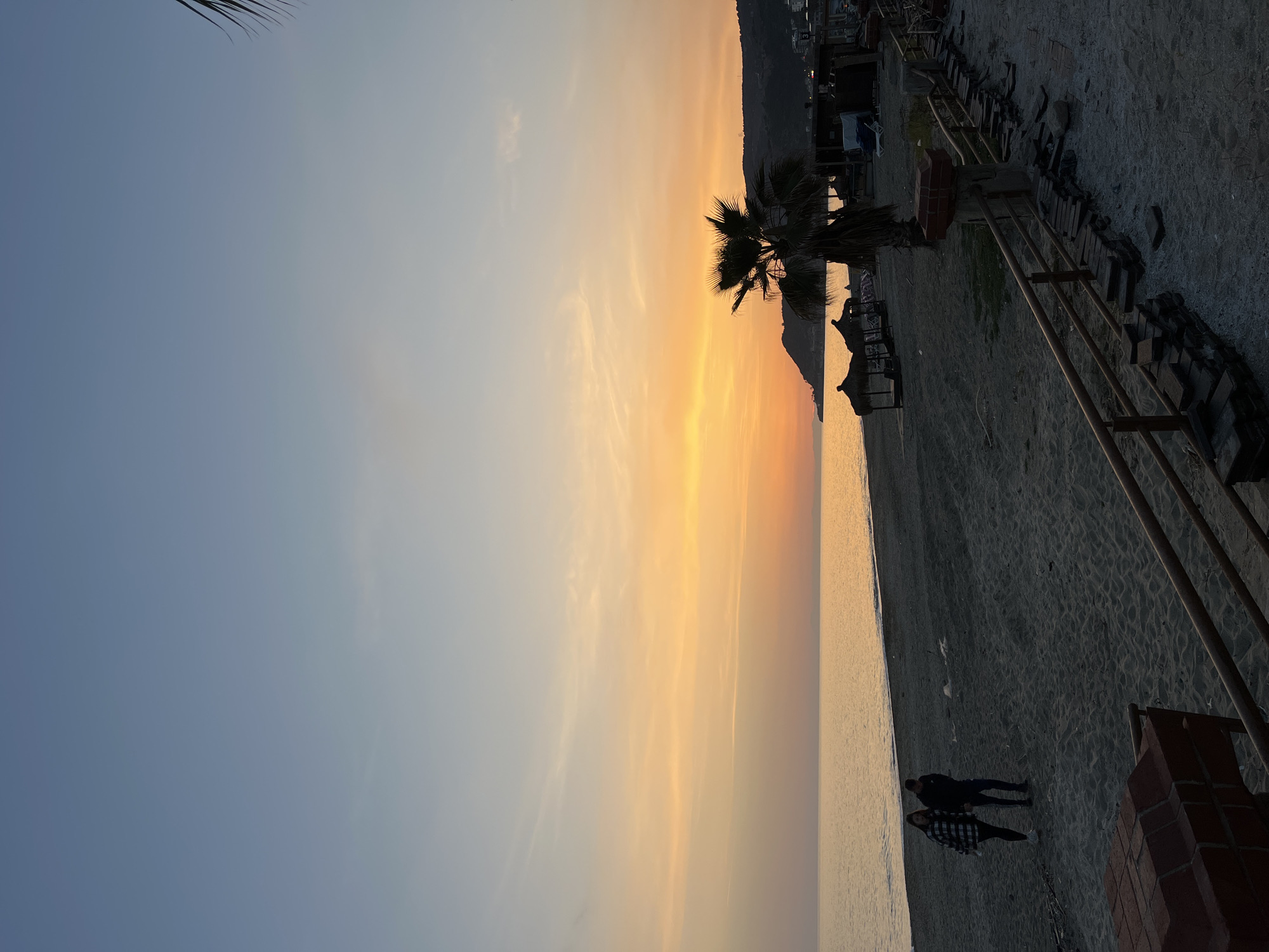 The image depicts a scene at Alanya, Turkey. The scene is a beach at sunset. There are two people walking on the sand near the water. A palm tree stands near a building with a thatched roof. There is a boardwalk or raised walkway next to the beach, constructed with brick and wood, and a metal railing. The sky displays a gradient of colors from blue to orange.