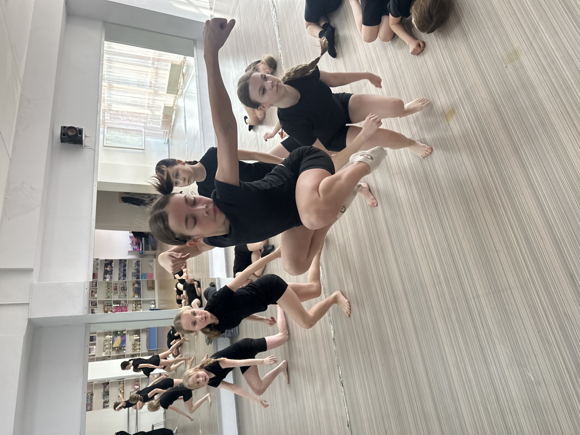 A group of young people, predominantly girls, are in a dance studio. They wear black dance attire. Some are barefoot while others wear ballet shoes. They are performing a dance routine, with arms raised and bodies in motion. The studio has a mirrored wall reflecting the dancers. A trophy shelf is visible behind the mirrored wall. The floor is light-colored with a striped pattern. A window and light fixtures are also present. The location is consistent with a dance studio or gymnasium located in an urban area. There is no indication of the city of Votkinsk, Russia being present in this image.