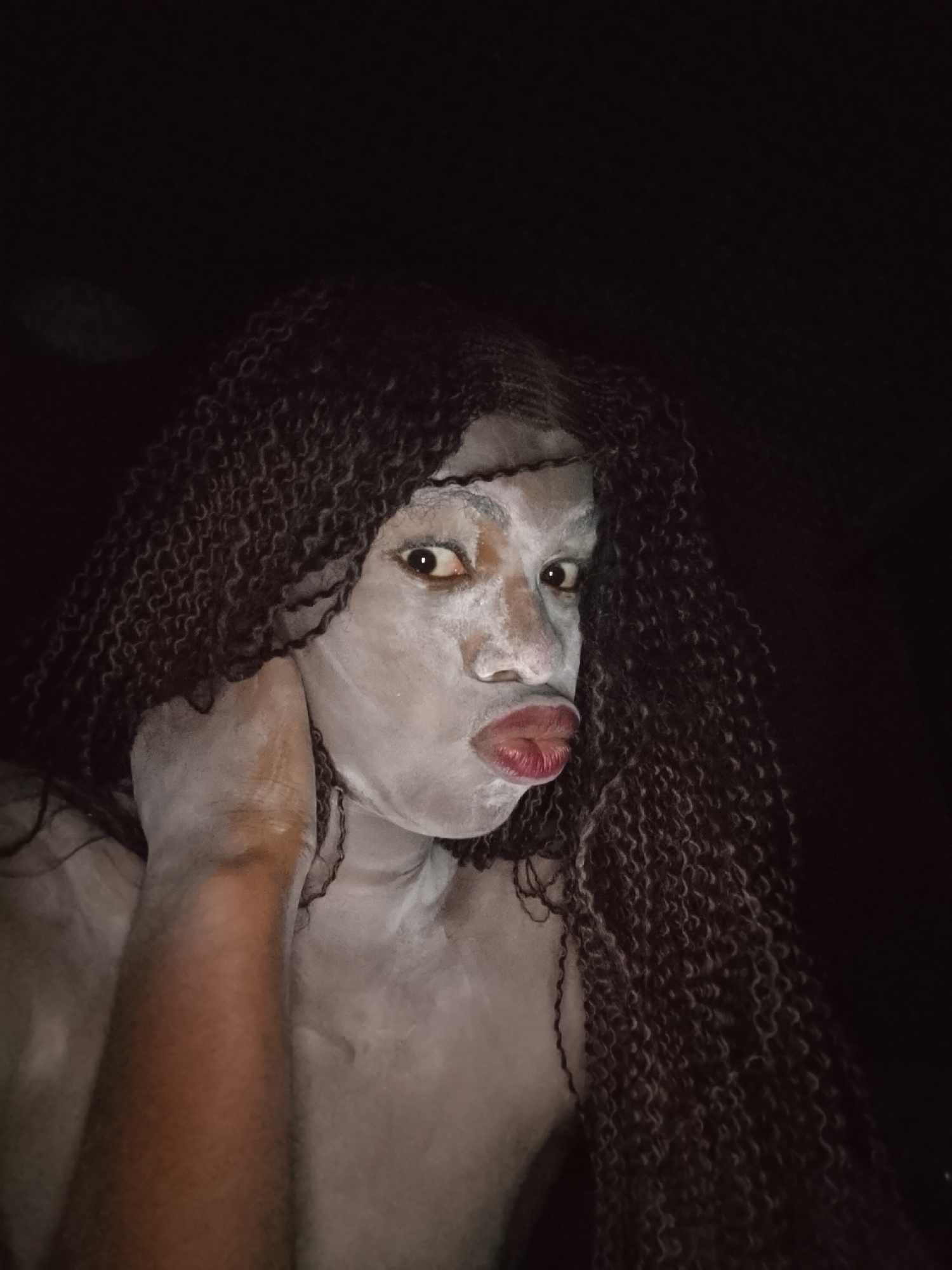 A dimly lit close-up photo shows a person with dark, curly hair wearing heavy white face paint, pouting their lips, and holding their neck. The person's chest is exposed. Due to the lack of landmarks, street signs, and specific cultural markers in the image, it is impossible to confirm if this picture was taken in Onitsha, Nigeria.