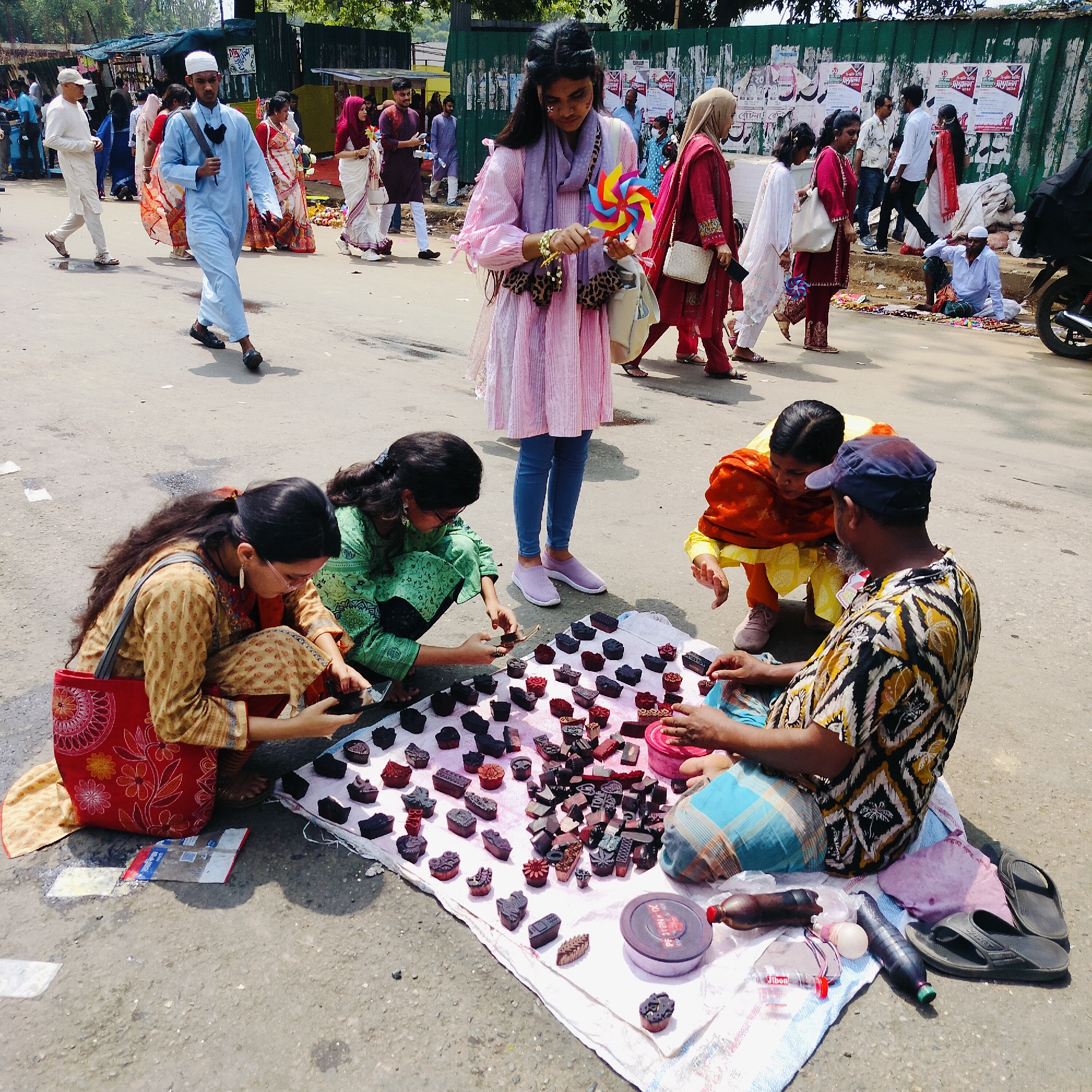 A medium shot on an overcast day depicts an outdoor market scene in Dhaka, Bangladesh. In the foreground, two young women crouch near a vendor, inspecting small, dark objects laid out on a white cloth spread on the asphalt. One woman, wearing a tan top and a red patterned bag, holds a dark object in her hand. The second woman wears a green outfit and glasses, also examining an object. The vendor, a dark-skinned man wearing a patterned shirt and a blue baseball cap, sits cross-legged, interacting with the women. A third woman, dressed in an orange and yellow outfit, also examines the merchandise.
Behind them, a young woman in a pink dress and blue jeans stands holding a colorful pinwheel. Numerous people walk along a street, and a man in a blue tunic stands out. Further back, crowds of people are visible, many wearing traditional Bangladeshi attire. The background shows a green fence and buildings, with posters displayed on the fence. A motorbike is partially visible on the right. The general atmosphere suggests a busy market day with people engaging in commerce and social interaction.