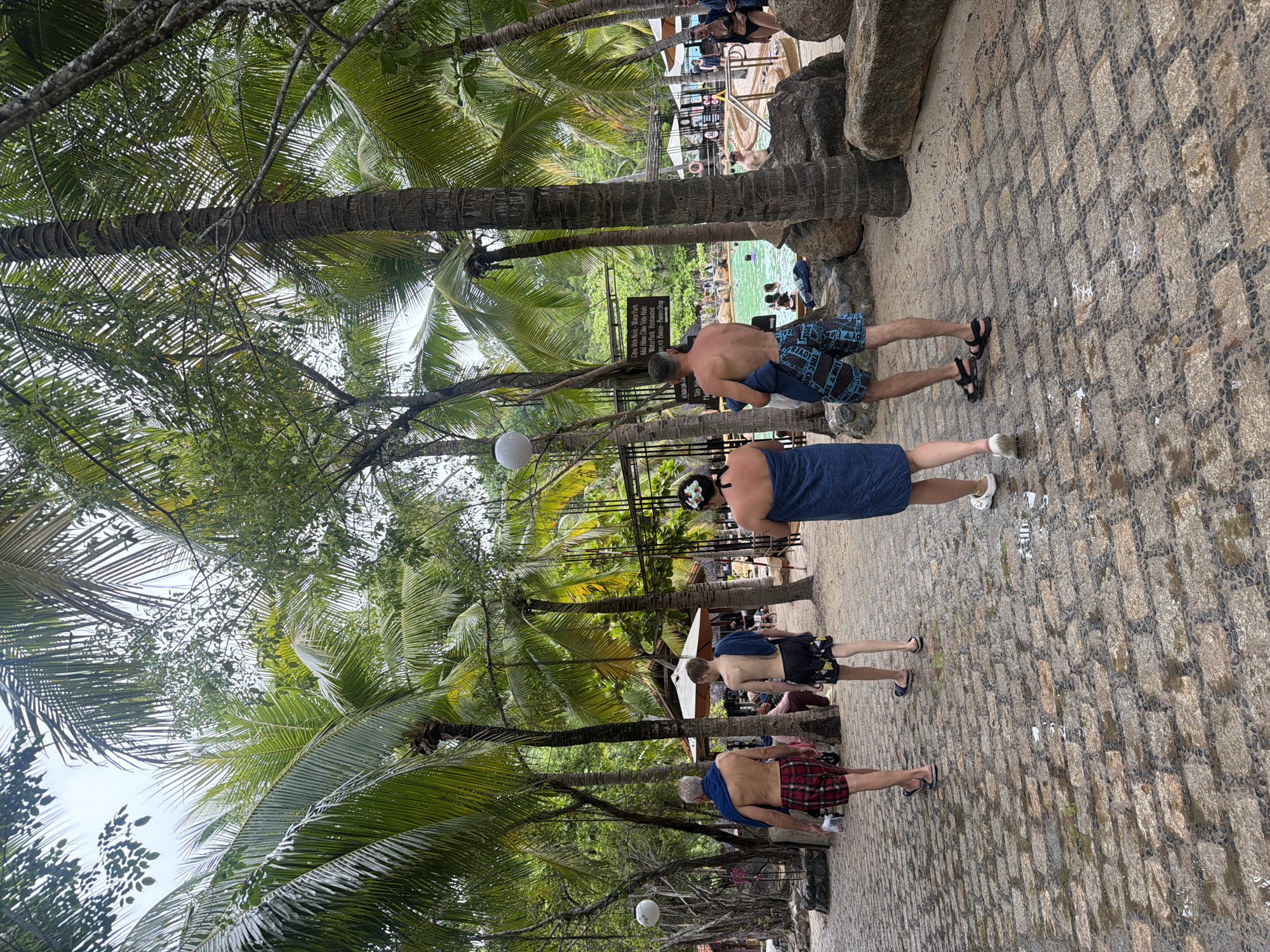 The image depicts a scene at a water park in Nha Trang, Vietnam, likely part of a resort complex, indicated by the presence of palm trees, a pool, and people in swimwear. In the foreground, four individuals are walking away from the camera on a wet, cobblestone pathway lined with large rocks and surrounded by lush vegetation, including numerous palm trees. Two men in the foreground are wearing plaid swim trunks and patterned swim shorts, respectively, and both have towels draped over their shoulders. A woman in the middle foreground is wrapped in a blue towel and wearing white sandals. Another man to her right is also wrapped in a blue towel and wearing patterned swim shorts and black sandals. In the background, a bright green pool is visible with several people swimming and relaxing. There are also lounge chairs, umbrellas, and other amenities associated with a water park or resort. The overall atmosphere suggests a tropical vacation destination.