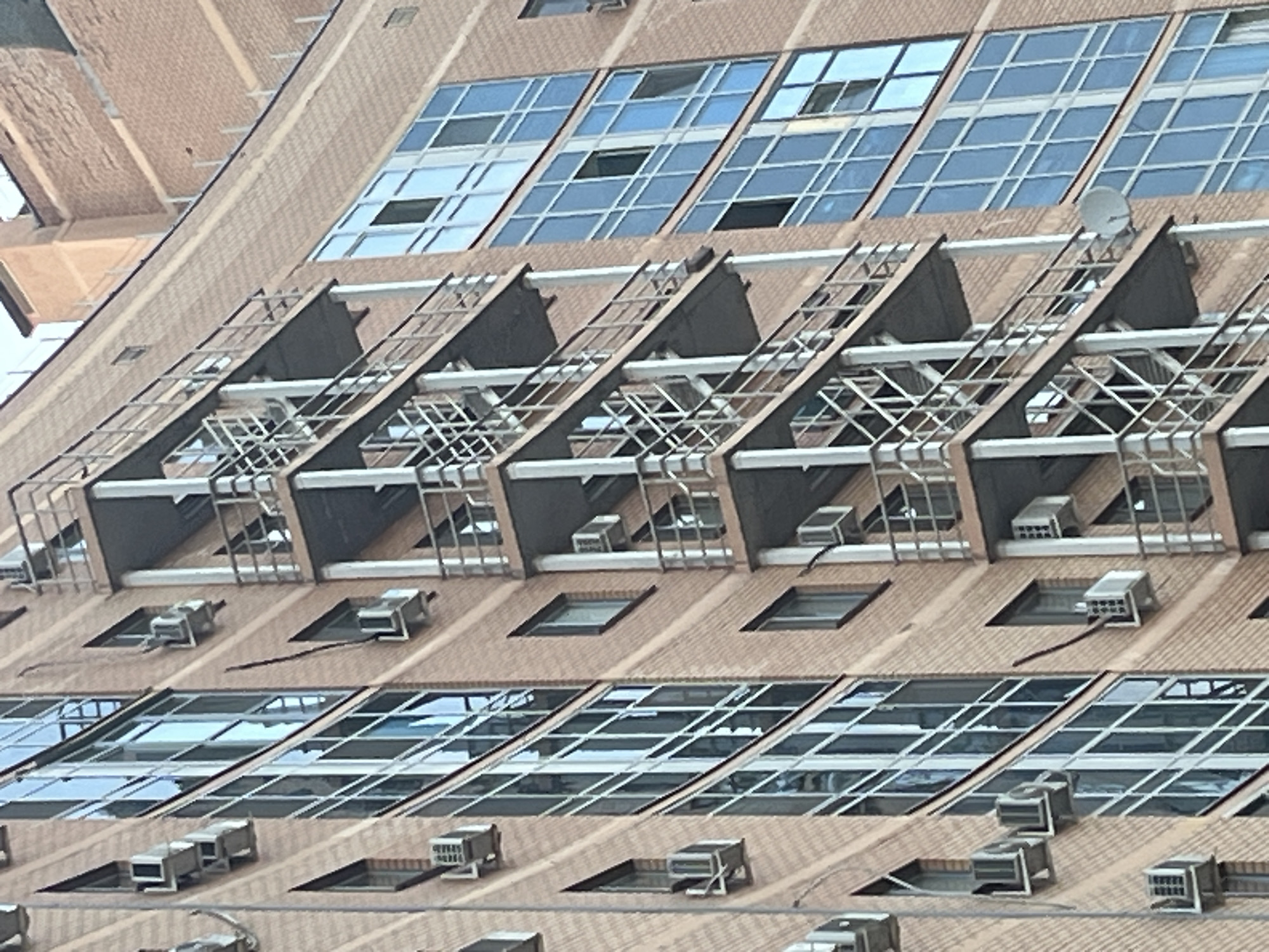 The image depicts the exterior of a building in an urban environment, specifically featuring a facade with a repetitive pattern of windows and air conditioning units. The building is constructed of brick, and the windows are rectangular with a multi-pane design. Above each row of windows, there is a series of black, angular structures with a metallic framework that appears to be part of the building's design or possibly a system of external sunshades. The air conditioning units are visible in recesses along the brickwork. A satellite dish is also mounted to the structure. The image does not contain any discernible people, actions, or interactions. The location is Kazan, Russia.