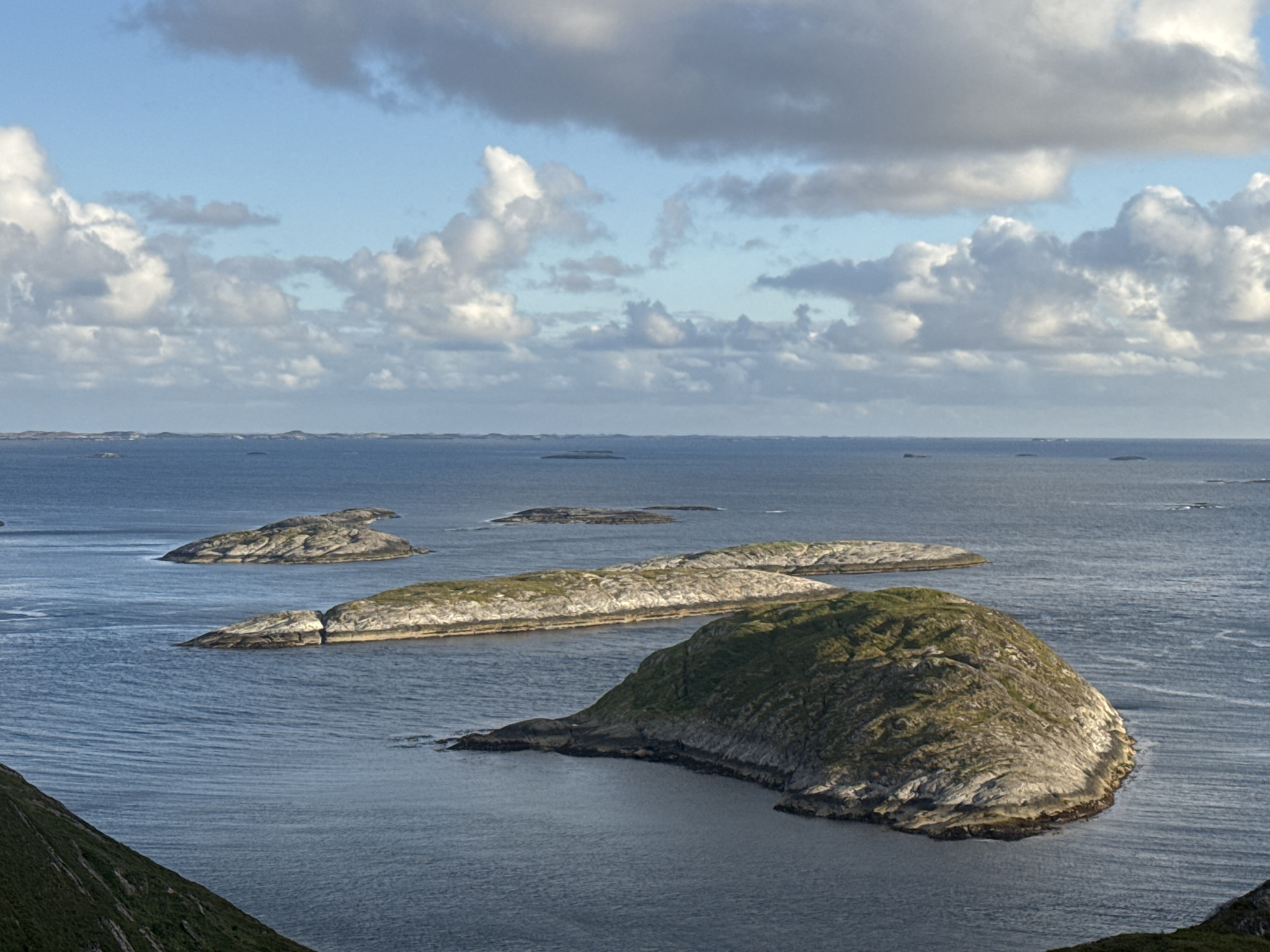 This is a scenic, wide-angle, eye-level photograph of the Sleneset archipelago in Norway, taken on a clear, partly cloudy day. The foreground features a steep, grassy hillside on the left, sloping down to dark, rippling water. In the middle ground, several rocky islands with patches of green vegetation dot the calm, blue sea. The islands vary in size and shape, with some appearing flat and others more rounded. In the background, the horizon is a thin line of more distant islands under a sky filled with scattered, white and grey clouds. The lighting suggests it is either late afternoon or early morning, with the sun casting soft light on the islands and highlighting the textures of the rocks and clouds. There are no people or animals visible in the image.