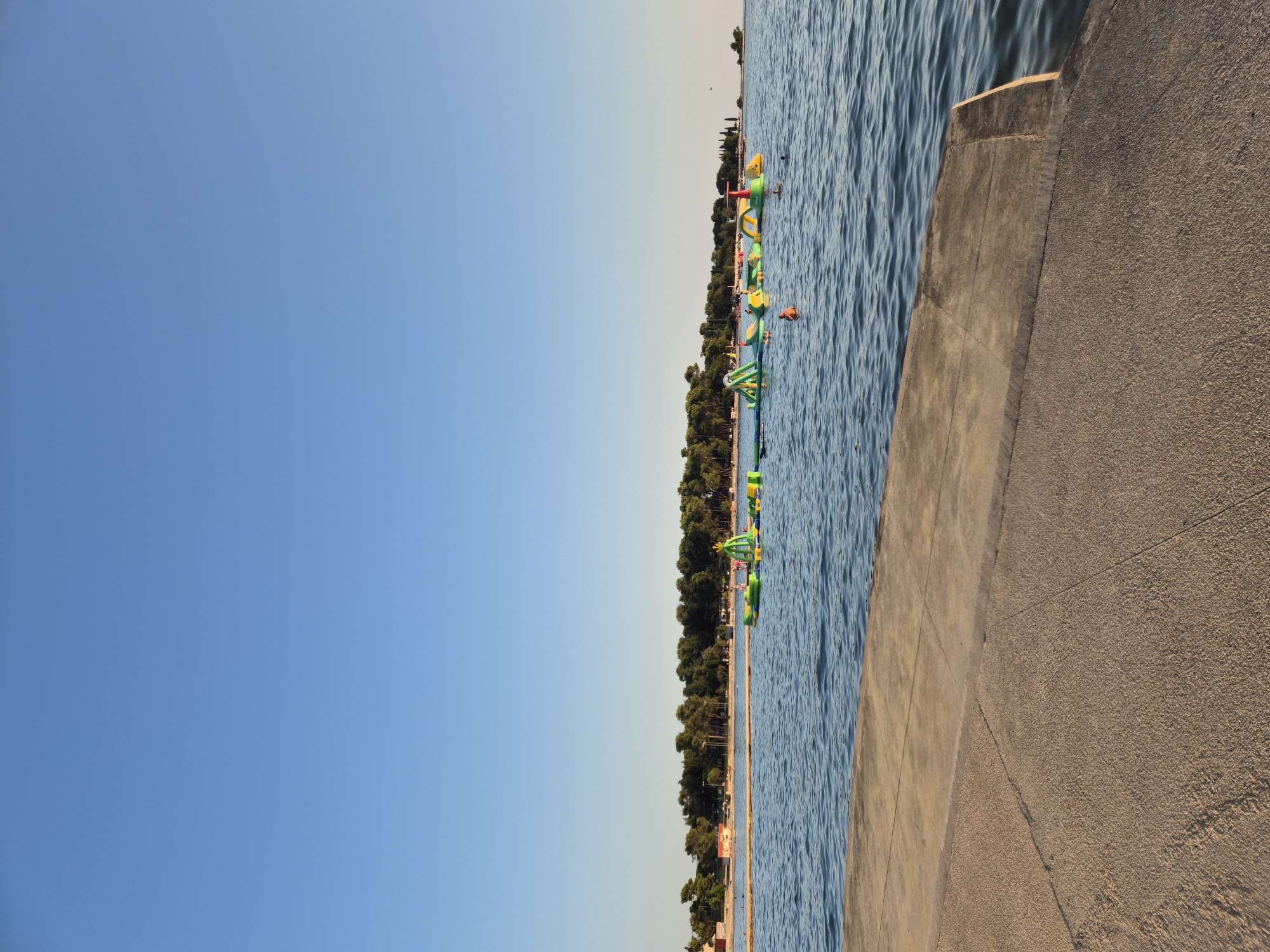 The image depicts a clear blue sky over the Adriatic Sea in Katoro, Croatia. In the foreground, a concrete pier or walkway extends into the water. In the middle ground, a vibrant inflatable water park is set up in the sea, featuring various green and yellow structures. Several people are visible in the water, some swimming and others interacting with the water park. The background shows a sandy beach lined with lush green trees, and beyond that, a small cluster of buildings. The overall scene is serene and suggests a recreational atmosphere on a sunny day.