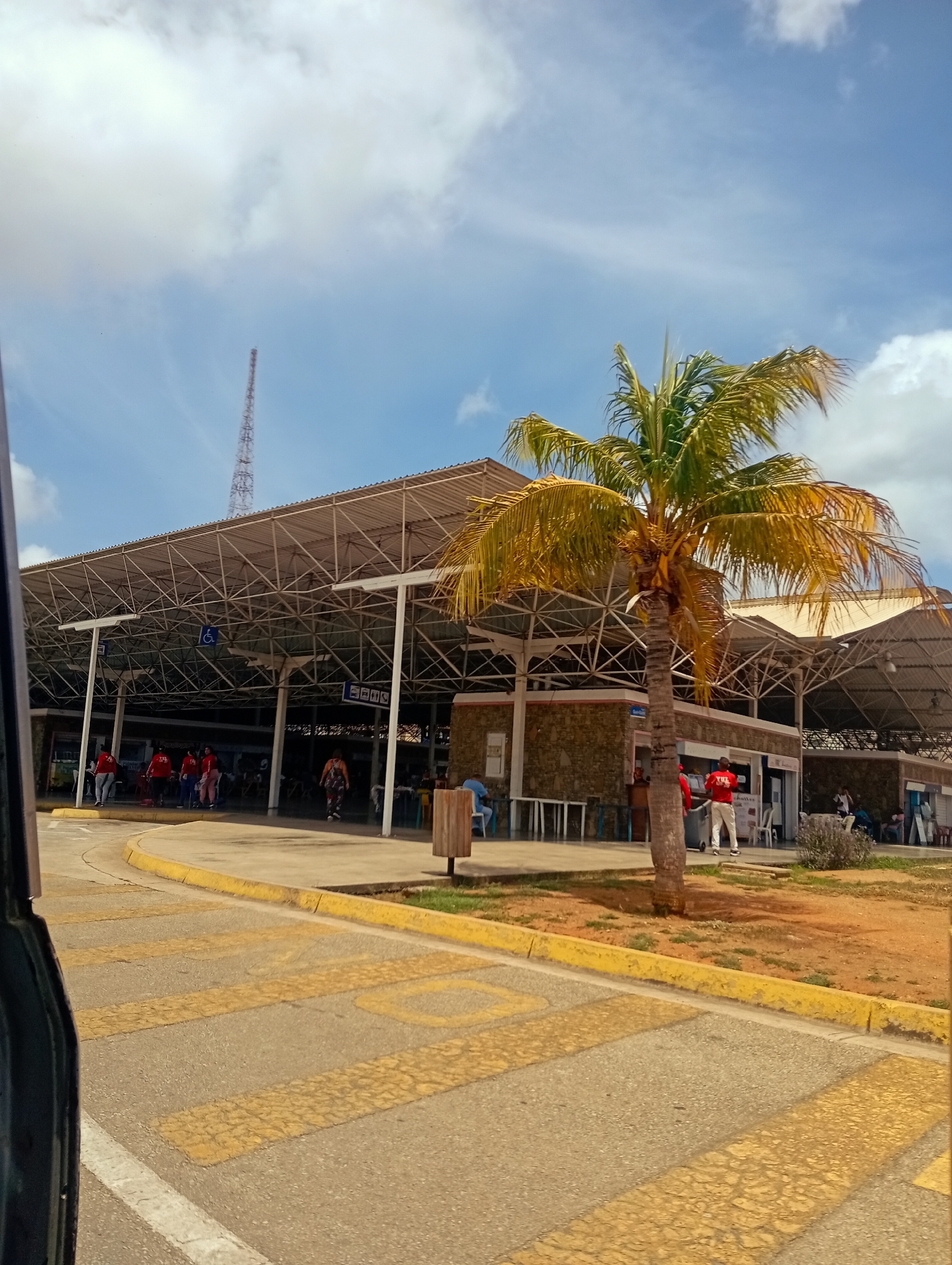 The image depicts the exterior of an open-air market or bus terminal in Juan Griego, Venezuela. A large, sloped roof supported by a metal frame covers an area where people are gathered. The facade of the building behind the roof is made of stone. Several individuals are visible in the foreground and midground, some standing and others appearing to walk. One person in the midground is wearing a red shirt and white pants, facing away from the camera. A tall palm tree with yellowing fronds stands to the right of the frame. The ground in front of the building is paved, with yellow markings on the asphalt, suggesting a roadway or parking area. The sky above is blue with scattered white clouds. A cell tower is visible in the distance, behind the structure. The overall impression is that of a public transportation hub or a bustling marketplace in a tropical or subtropical climate.