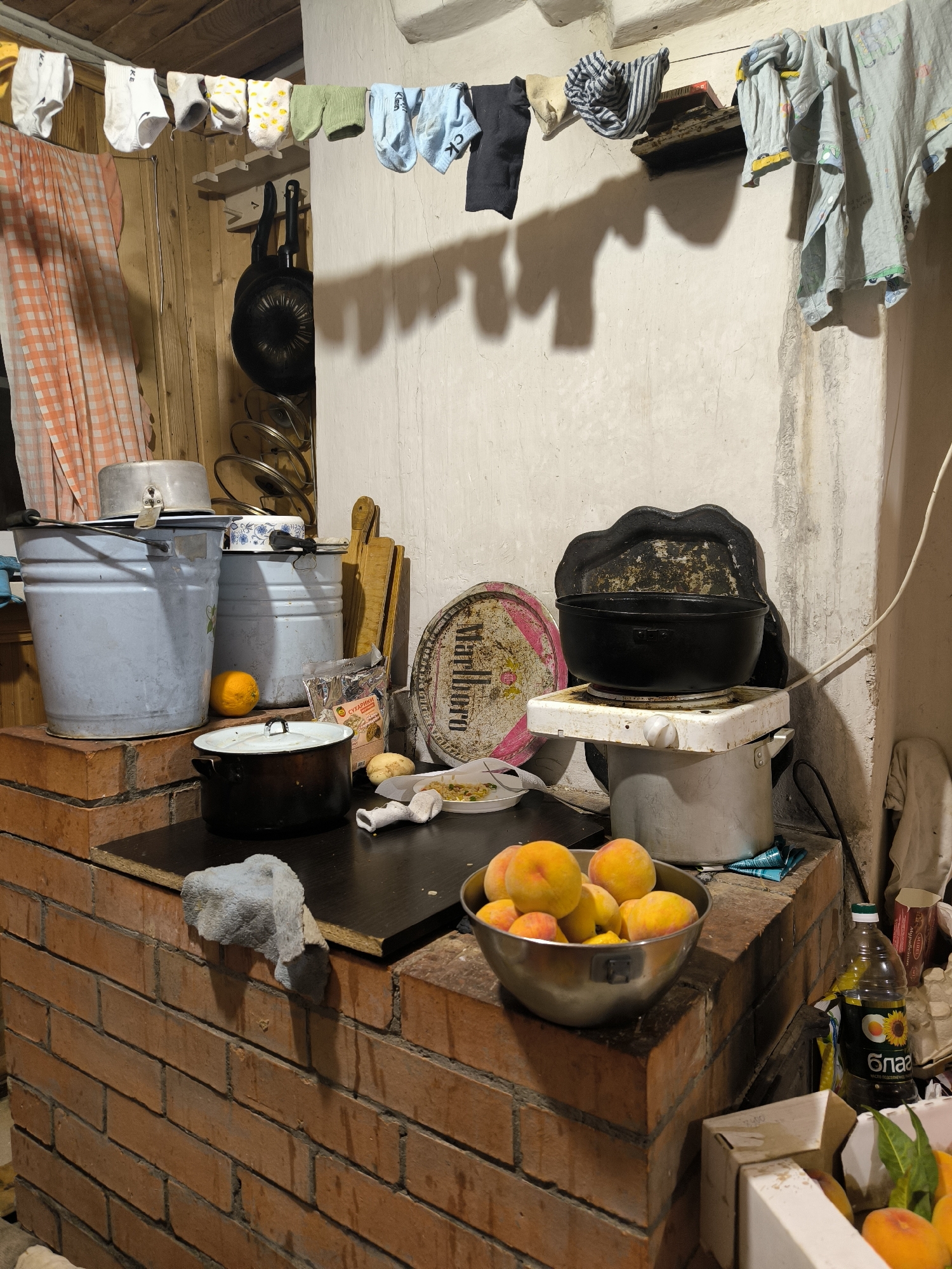 The image depicts a dimly lit, cluttered domestic interior, likely a kitchen area, featuring a brick stove. Laundry, including socks and garments, is hanging on a line above the stove. Various kitchenware is visible, such as pots, pans, buckets, and cutting boards. A small electric hot plate is topped with a black pot. A bowl of peaches is positioned on the counter. The environment appears rustic and somewhat unkempt, with a mixture of cooking implements, food items, and everyday household objects. The overall impression is one of a functional, albeit somewhat disorganized, living space. There are no people visible in the image. The image's context implies it is located in Uzunovo, Russia.