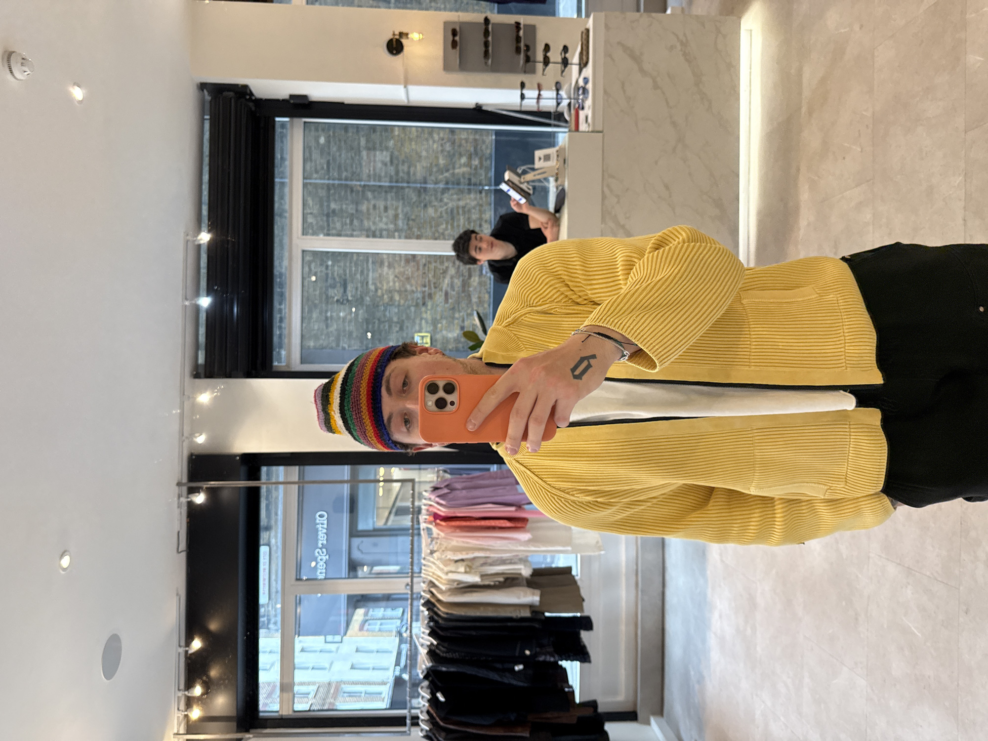 This image captures a mirror selfie taken inside a modern retail store or boutique. The main subject, seen from the chest up, is a young person wearing a multi-colored knitted beanie (featuring rainbow stripes) and a bright yellow, horizontally ribbed long-sleeved top. They hold an orange-cased smartphone, through which the photo is taken, and look directly at the camera with a neutral expression. A distinct angular tattoo is visible on their left forearm.
In the background, another person with dark hair, dressed in a black top, is seated behind a light-colored, marble-patterned counter, engrossed in a phone or tablet. Behind them, large windows overlook an urban street, revealing brick buildings, consistent with the given London, United Kingdom context.
The store's interior is brightly lit with recessed and track lighting, featuring clean white walls and a light-toned, marble-patterned floor. Retail displays include racks of folded clothing in various colors (purple, pink, white, beige, grey, black) and shelves showcasing sunglasses or glasses on a back wall. Reflected on a window or sign, the reversed text "Olivier Spen" is visible, possibly a brand or store name. The lighting suggests it is daytime.