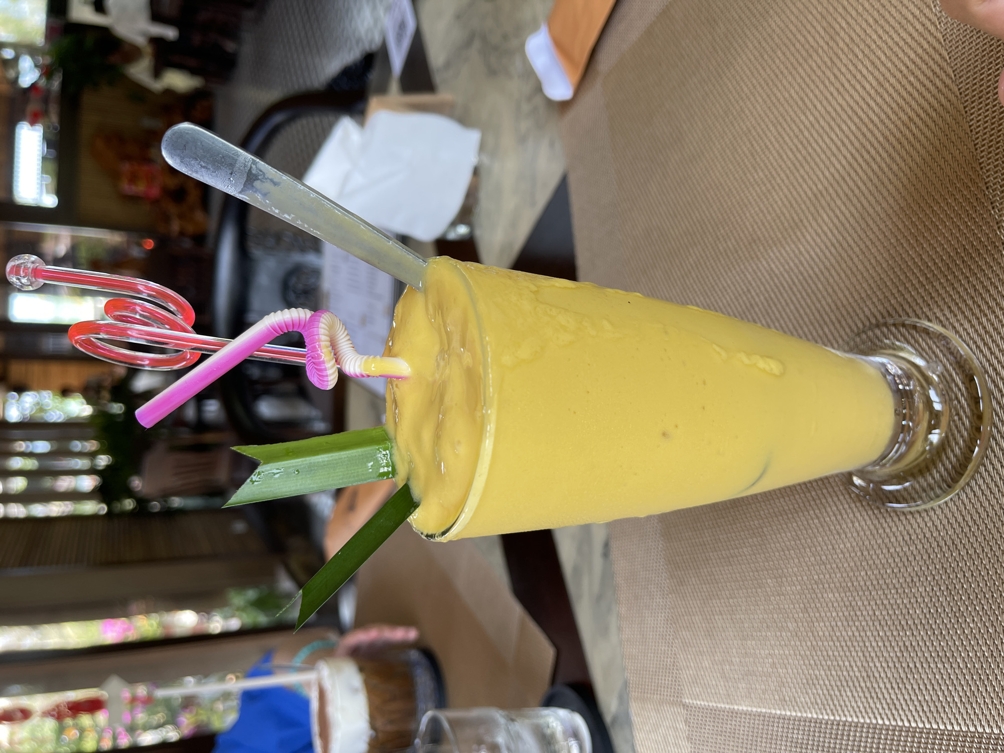 The image captures a vibrant yellow, thick beverage, likely a fruit smoothie or shake, served in a tall, clear glass with a stemmed base. The drink appears rich and creamy, with a few small specks suggesting it's made from fresh fruit, possibly mango, common in tropical locations like Phú Quốc, Vietnam.
Three straws are present: a clear, broad plastic spoon-straw partially submerged in the drink, a pink and white striped flexible straw also in the beverage, and a decorative red curly straw standing upright beside them. A fresh green leafy garnish, possibly a pandan leaf, is elegantly tucked into the side of the glass, adding a natural and aesthetic touch.
The glass rests on a light brown placemat with a distinctive diagonal woven pattern. The background is softly blurred, revealing a relaxed indoor or semi-outdoor dining setting. Hints of dark furniture, possibly rattan chairs, and ambient lighting suggest a daytime scene at a restaurant or cafe in Phú Quốc. The overall impression is one of a refreshing and exotic drink ready to be enjoyed in a pleasant, vacation-like atmosphere. No people or visible text are present in the image.