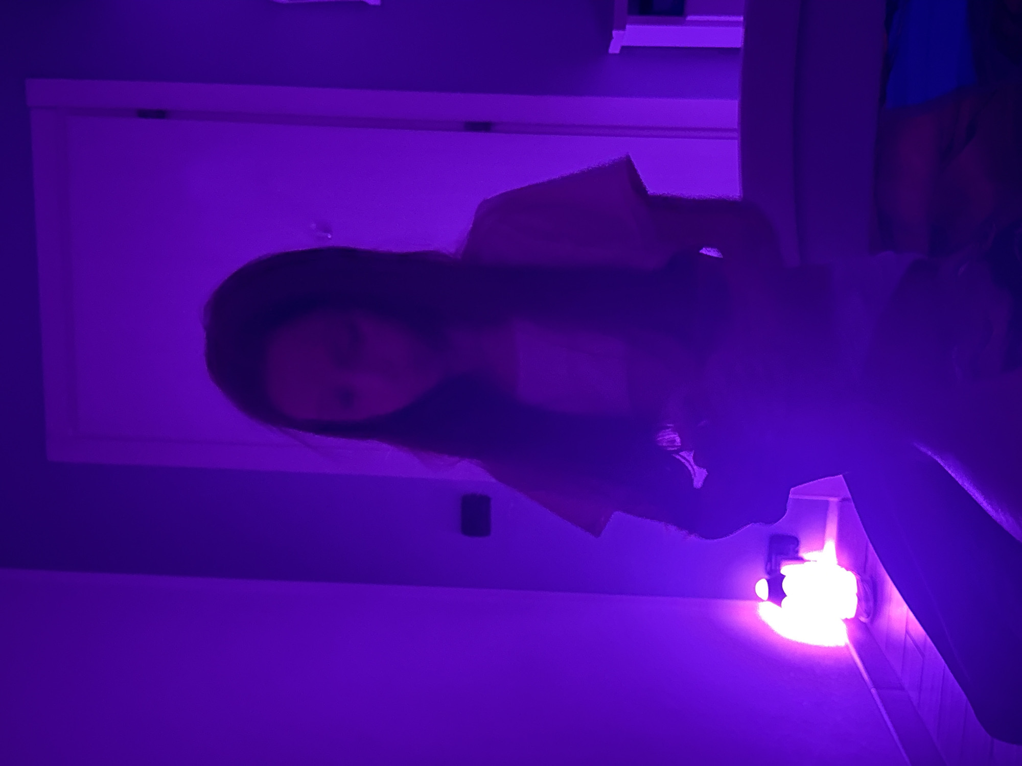 In a room illuminated by purple light, a young girl with long, dark hair sits on what appears to be a bed. She is looking down at something in her hands, partially obscured by the low light and her posture. To the left, a bright, white lantern sits on a surface, casting a strong glow that mixes with the ambient purple light. The background reveals a closed white door and a section of wall. The overall impression is one of a softly lit, personal space. The image does not contain any discernible elements that can place it specifically within the city of Genoa, Italy.