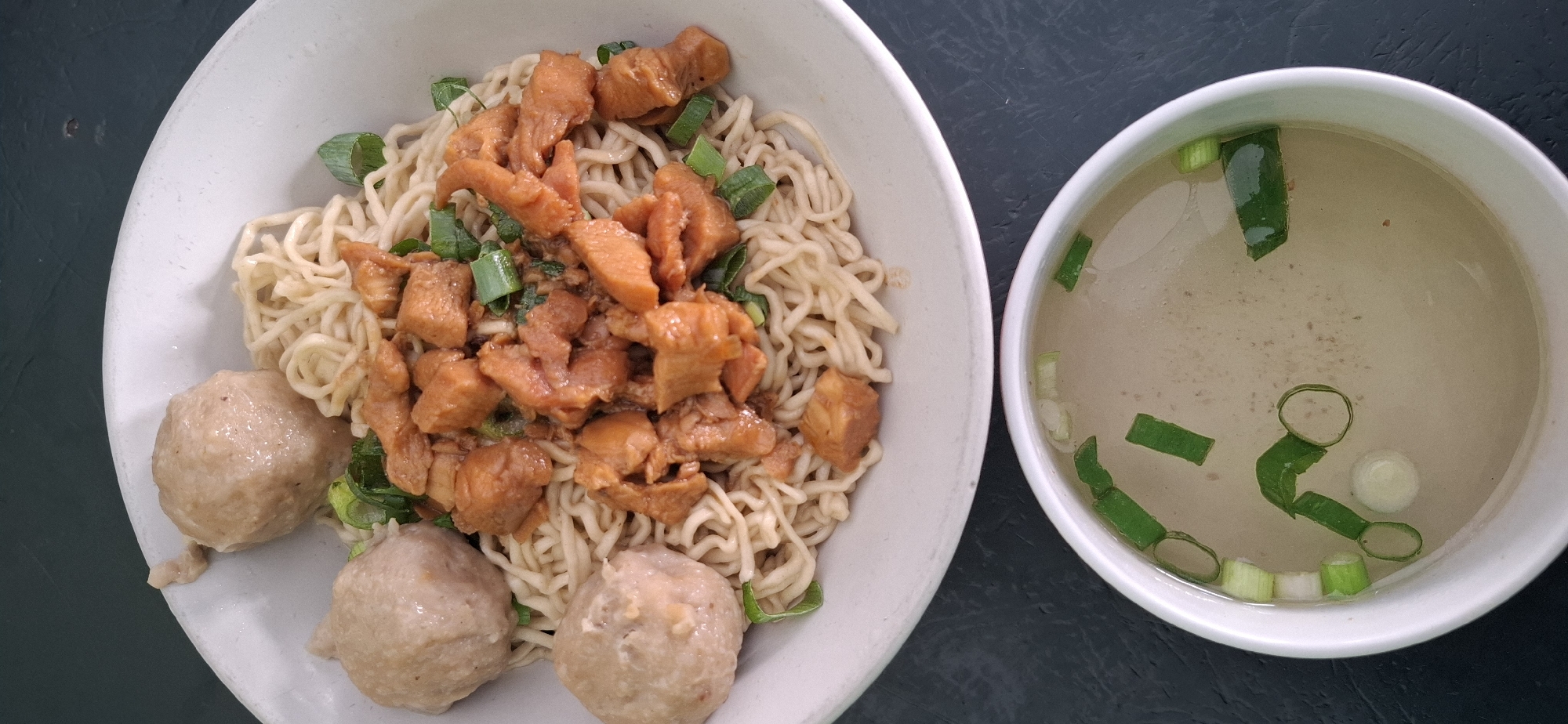 A close-up, high-angle shot shows a bowl of noodles topped with diced chicken and green onions, served with a small bowl of broth garnished with more green onions. The noodles are thin and pale yellow, intertwined with pieces of browned, cooked chicken. Chopped green onions are scattered throughout the noodles and on top of the chicken. To the left of the noodle bowl are three round meatballs. The broth in the smaller bowl is a light, translucent yellow and contains chopped green onions and some small specks, possibly sesame seeds. The dishes are presented on a dark, textured surface, which could be a table. This image captures a typical serving of "Mie Ayam" (chicken noodles), a popular street food dish in Indonesia, likely photographed in a restaurant or eatery in Kartasura, Indonesia.