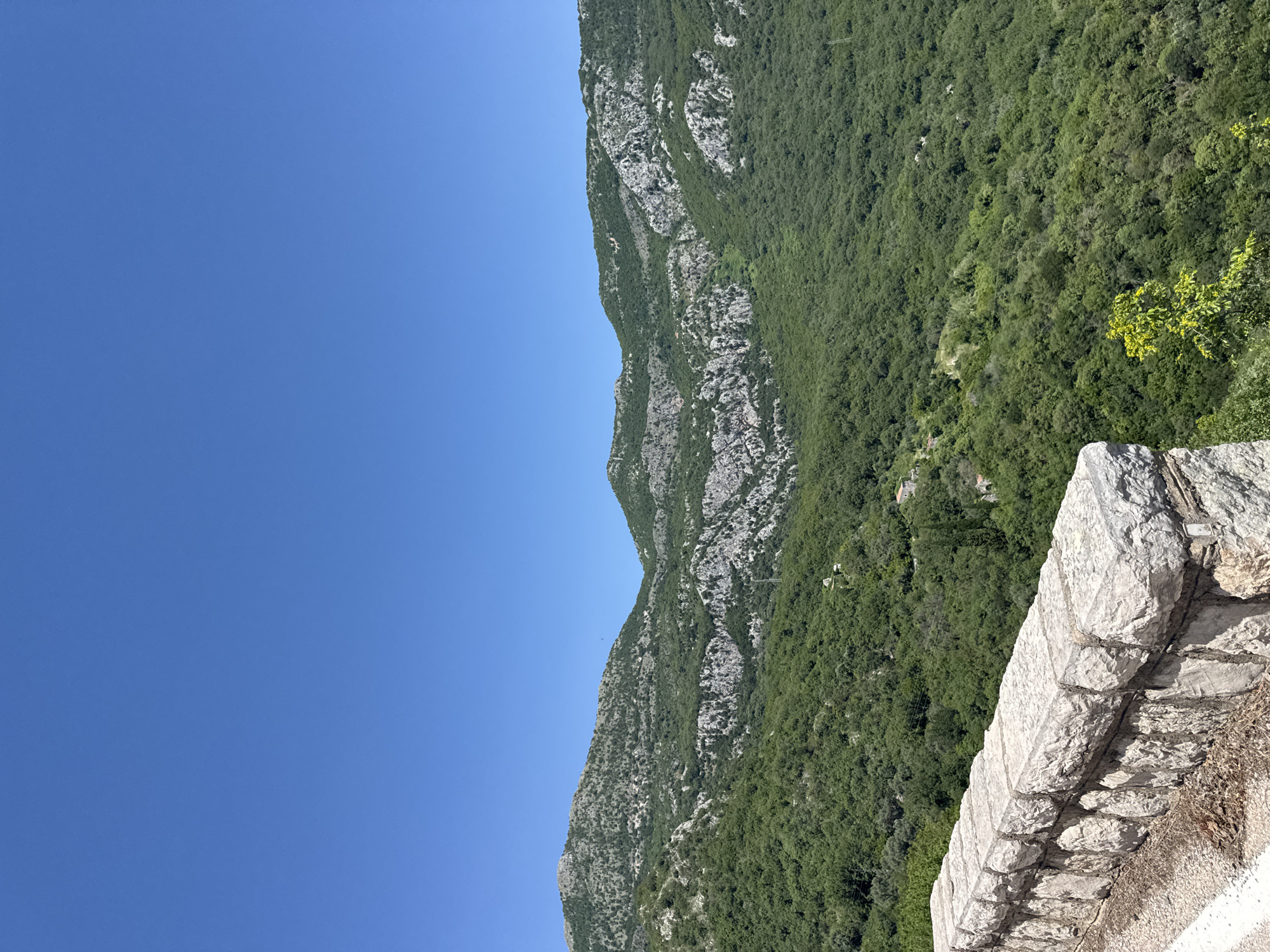 The image captures a scenic view from Petrovac na Moru, Montenegro. The foreground features a stone wall, likely a part of a viewing platform or a building's structure. Beyond the wall, a steep mountainside is covered with lush green vegetation, interspersed with rocky outcrops and cliffs. The mountain ridge ascends towards the sky, forming a dramatic backdrop. Above the mountain, the clear blue sky suggests a sunny day. The photograph is taken from a slightly elevated position, giving a panoramic perspective of the natural landscape.