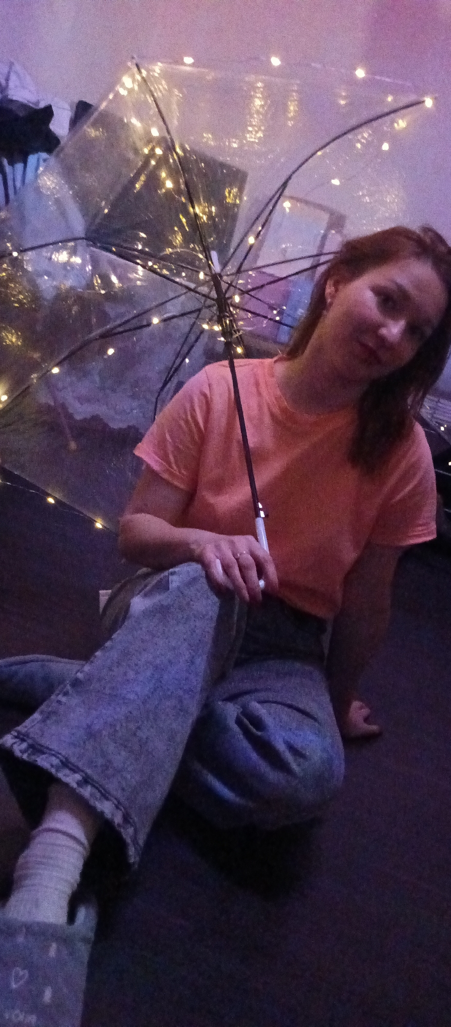 A young person, likely female, with medium-length reddish-brown hair, sits on a dark floor in an indoor setting. She is positioned towards the left of the frame, looking directly at the camera with a soft, slightly smiling expression.
She is holding a transparent umbrella embedded with numerous small, glowing fairy lights, which provide a significant portion of the illumination in the dimly lit scene. The umbrella's ribs are visible through the clear material.
The person is wearing a short-sleeved, plain orange or peach-colored t-shirt, light blue or grey acid-wash wide-leg jeans, white ribbed socks, and grey slippers with a faint heart pattern visible on the toe. Her right hand is holding the handle of the umbrella, with a ring visible on one finger.
The background is blurry and dimly lit with purple and pinkish hues, suggesting other objects or furniture, possibly boxes or reflective surfaces, creating a soft, ethereal ambiance. The overall impression is one of a cozy, whimsical moment, likely captured in the evening or at night, highlighted by the glowing umbrella. No discernible text is visible.