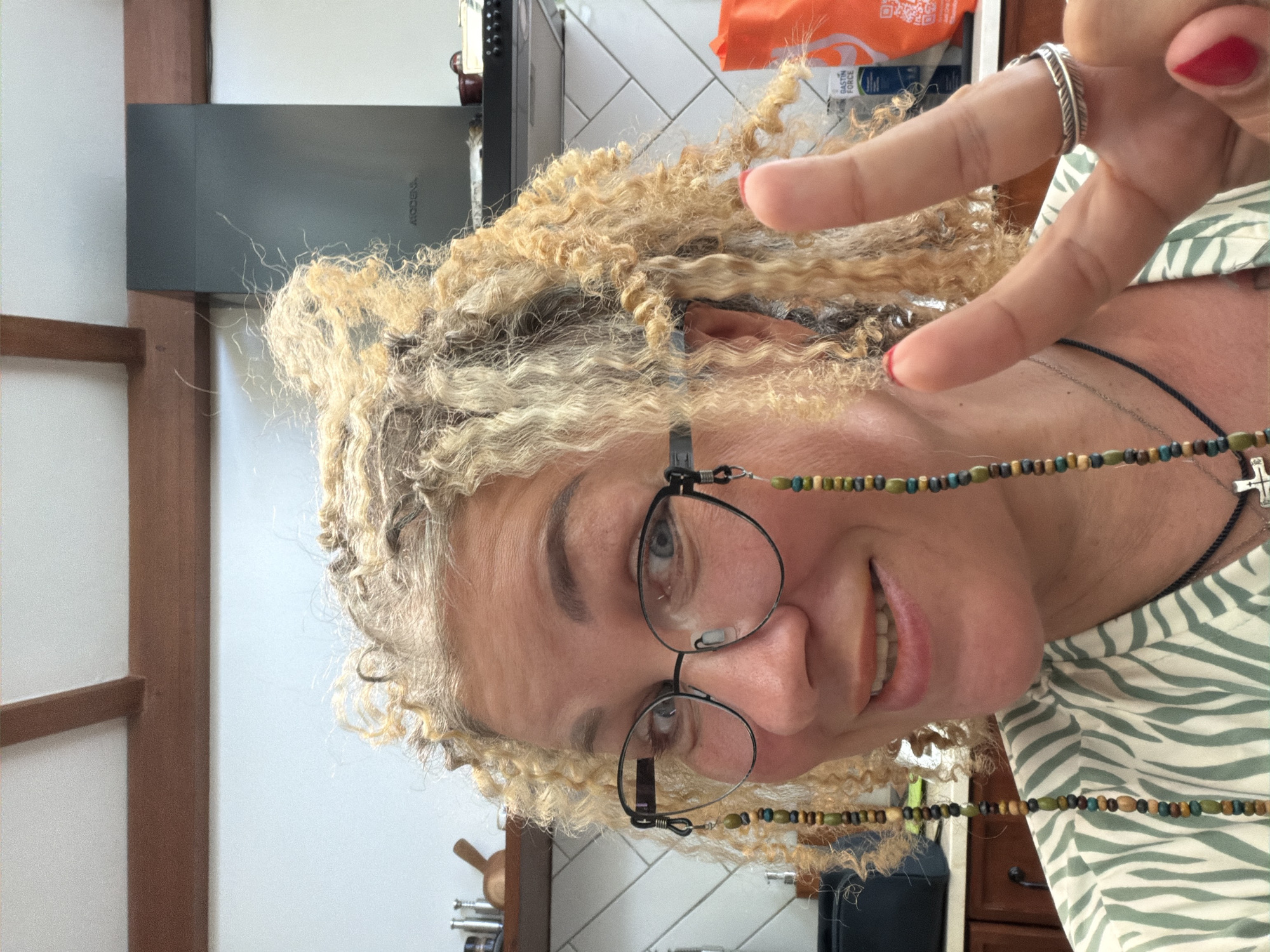 A person with blonde curly hair is wearing glasses with a beaded chain and making a peace sign with their right hand. They have a silver ring on their index finger and red nail polish. They are wearing a striped shirt and a cross necklace. The background shows a kitchen with white tiled walls, wooden cabinets, and a black appliance. The location is Unknown, Unknown.