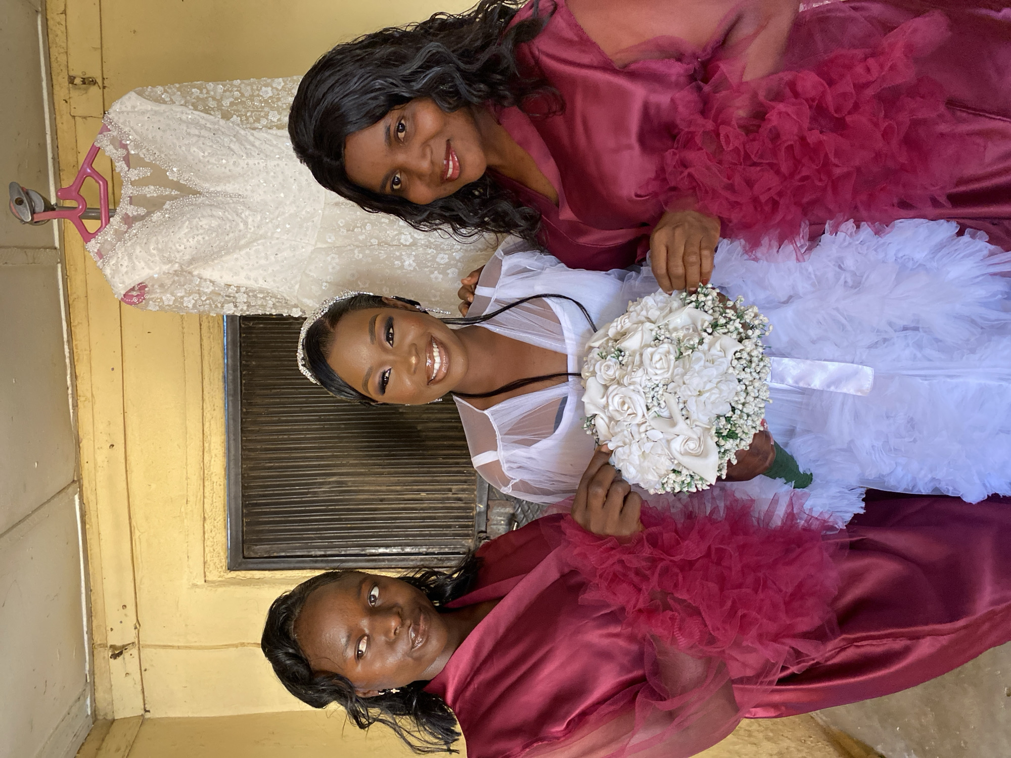 Three African women are posing for a photograph. One woman, the bride, wears a white dress and holds a bouquet of white flowers. The other two women wear maroon robes with ruffled sleeves. The bride has a jeweled headband. A beaded white dress hangs in the background. The photograph appears to be taken outside of a building in Vom, Nigeria. The wall behind them is painted yellow.
