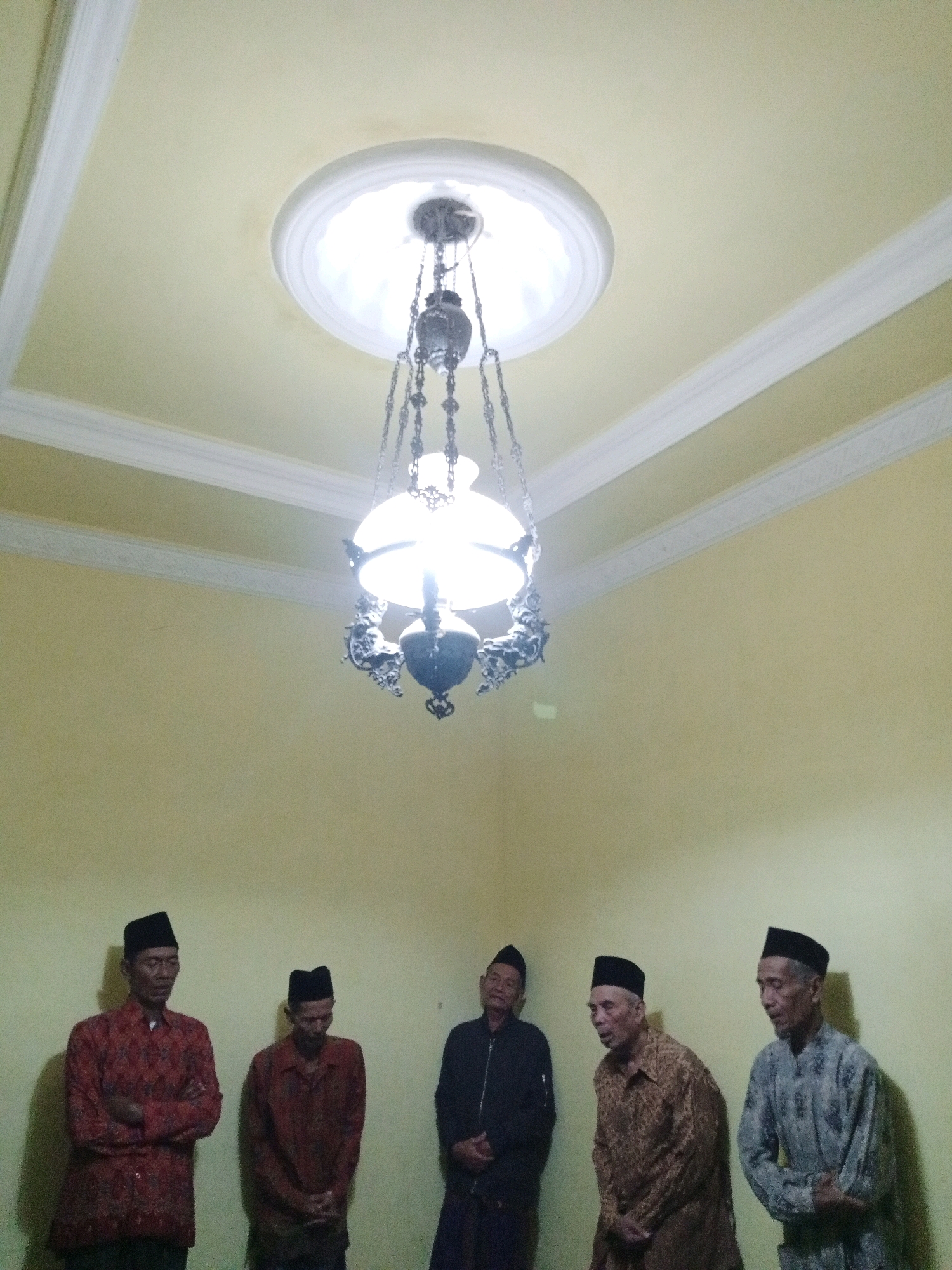 In the city of Sumberkepuh, Indonesia, five adult males are positioned indoors, standing against a pale yellow wall. They are all wearing traditional Islamic attire, specifically peci hats and long-sleeved shirts or jackets, with three of them also wearing sarongs. The individuals are standing in a line from left to right. The leftmost man is wearing a red and black patterned shirt. Next to him, another man is wearing a dark red shirt. The third man from the left is wearing a black jacket. The fourth man is dressed in a brown patterned shirt, and the man on the far right is wearing a grey patterned shirt. Above them, a vintage-style chandelier hangs from a decorative ceiling. The chandelier has a glass shade and intricate metal detailing. The ceiling itself is yellow with white molding. The lighting in the room appears to be provided by the chandelier.