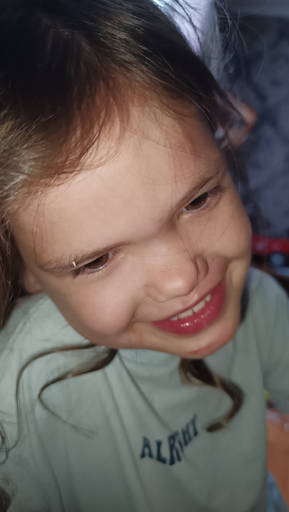The image is a close-up, high-angle shot of a young girl's face, focused on her eyes and mouth. Her head is tilted to the left, and her gaze is directed downwards and to her right. She has dark brown hair, some of which is falling around her face. Her eyes are brown, and she has a wide, shiny smile, revealing her teeth. She is wearing a light-colored t-shirt with dark blue lettering that appears to say "ALRHYT". The background is out of focus and appears to be an indoor setting with some fabric textures visible. The image does not provide any information about the city of Karsha, Russia.