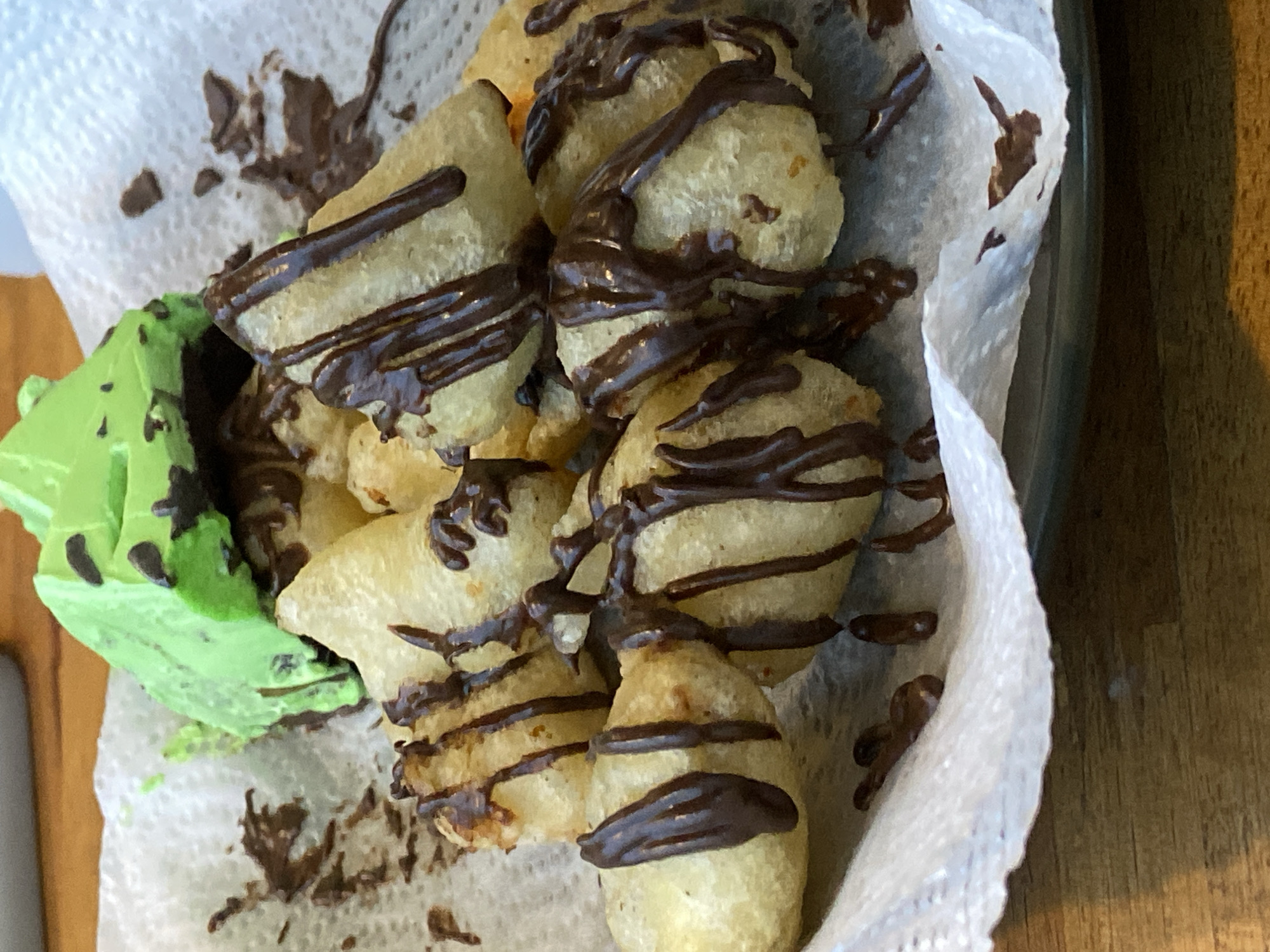 A pile of golden fried pastry pieces, drizzled with dark chocolate sauce, sits on a white paper napkin. A bright green, possibly lime-flavored, dessert or ice cream scoop is placed on top of the pastries. The food is arranged on a dark plate, which is on a wooden table. This scene is located in Candirejo, Indonesia.