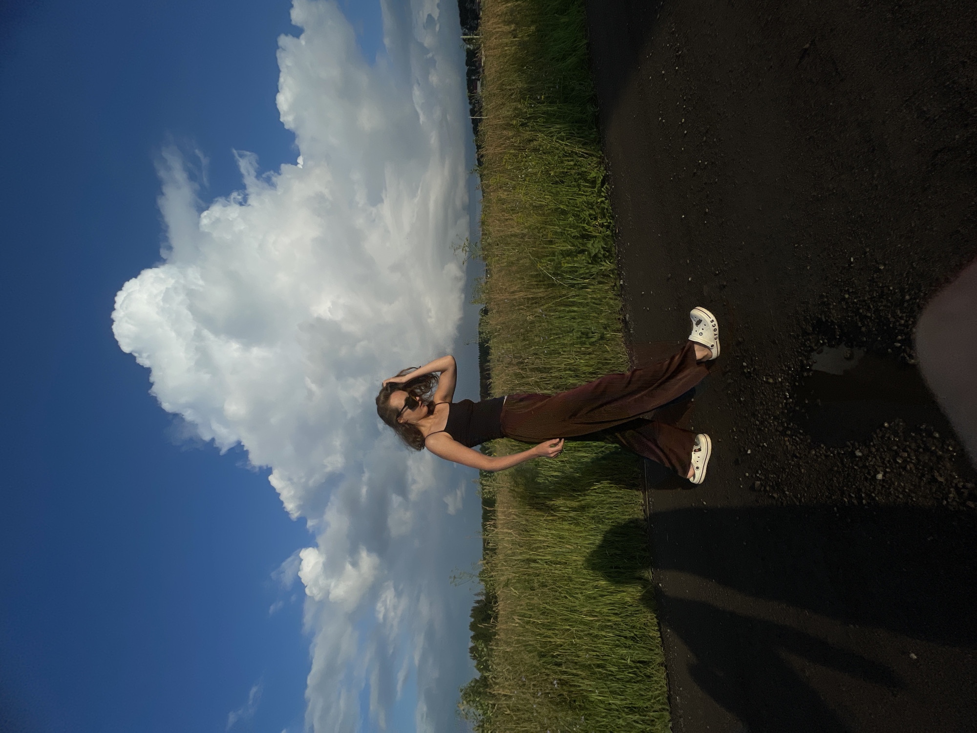 A young woman walks along a dark, paved path, her shadow stretching out to her left. She wears a black tank top, brown wide-leg pants, sunglasses, and white Crocs. She is in a grassy field under a bright blue sky with large, dramatic white clouds. In the distance, a line of trees and buildings is visible. The setting appears to be a rural area, potentially on the outskirts of a village like Podshivalovo, Russia, given the vast open space and agricultural landscape. The lighting suggests it is late afternoon or early evening, with the sun casting long shadows.
