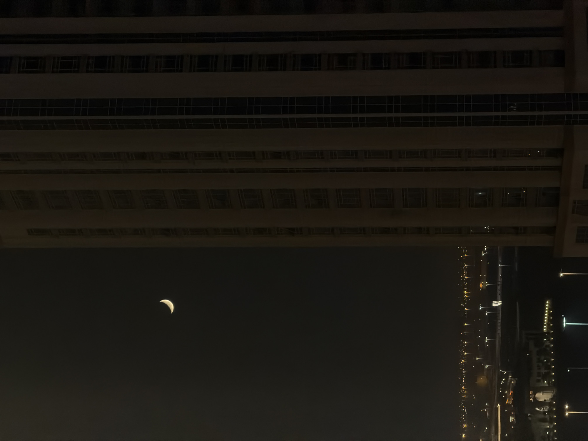 A crescent moon is visible in the dark sky to the left of a tall, modern building. The building's facade is light-colored with vertical dark window frames. Below the moon, a city skyline is illuminated by numerous lights, suggesting it is nighttime. The cityscape includes buildings of various sizes and streetlights. The overall scene is dark and atmospheric, with the moon and city lights providing the primary sources of illumination.