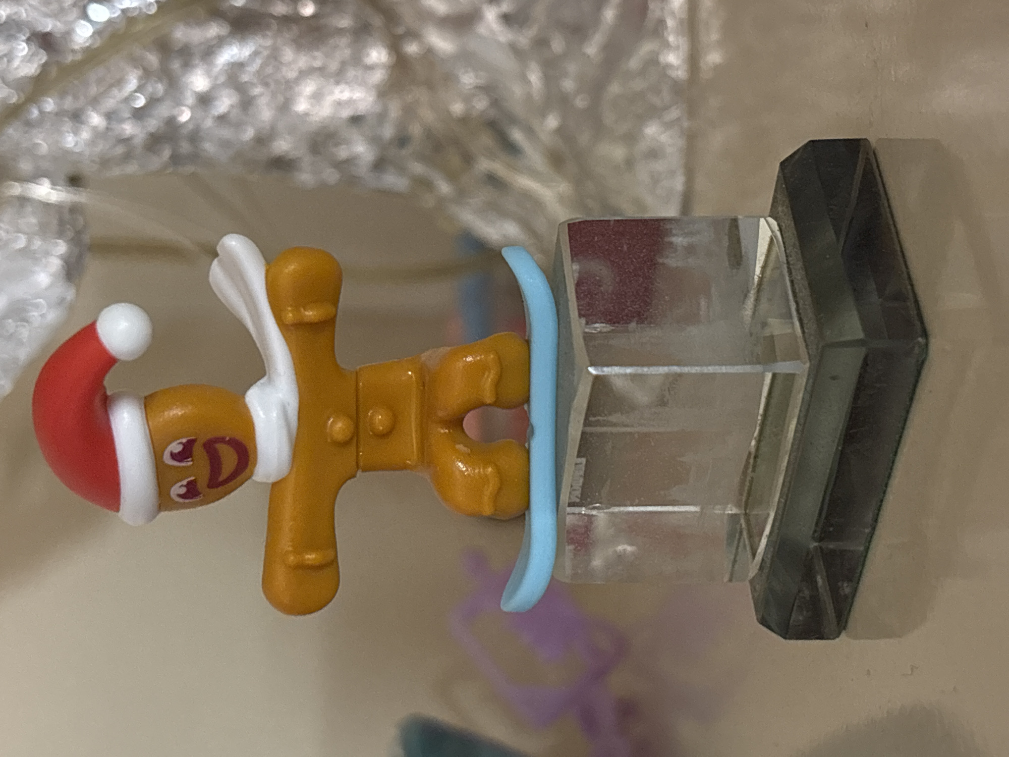 A toy gingerbread man wearing a Santa hat and white wings is attached to a clear, multi-sectioned prism stand with a black hexagonal base. The toy appears to be photographed on a reflective surface. The background is blurred and indistinct. There is no evidence to suggest the location is Sofia, Bulgaria.