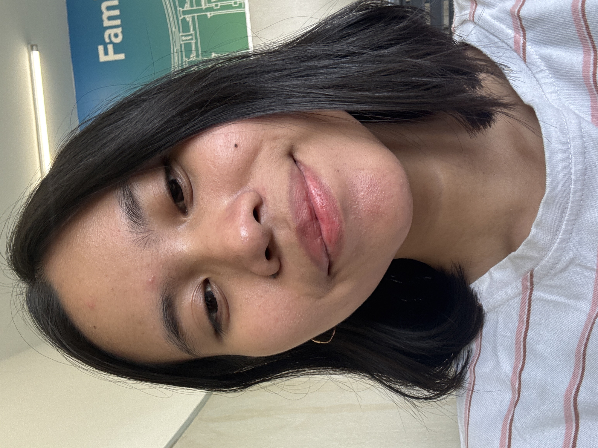 A young woman with dark hair and a light complexion smiles at the camera. She is wearing a white t-shirt with pink and brown stripes. A faint mole is visible on her right cheek. In the background, a blue sign with the word "Family" is partially visible, along with a fluorescent light fixture. The overall impression is that of a casual selfie taken indoors, possibly in an office or public building.